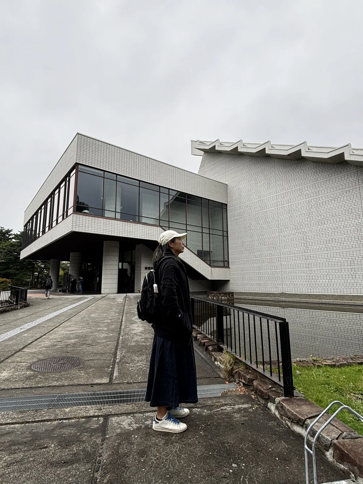 入口前的廣場，左側為懸挑的玻璃量體與室外樓梯，右側為白色磁磚的主體量體與階梯狀簷口，地面鋪設磁磚