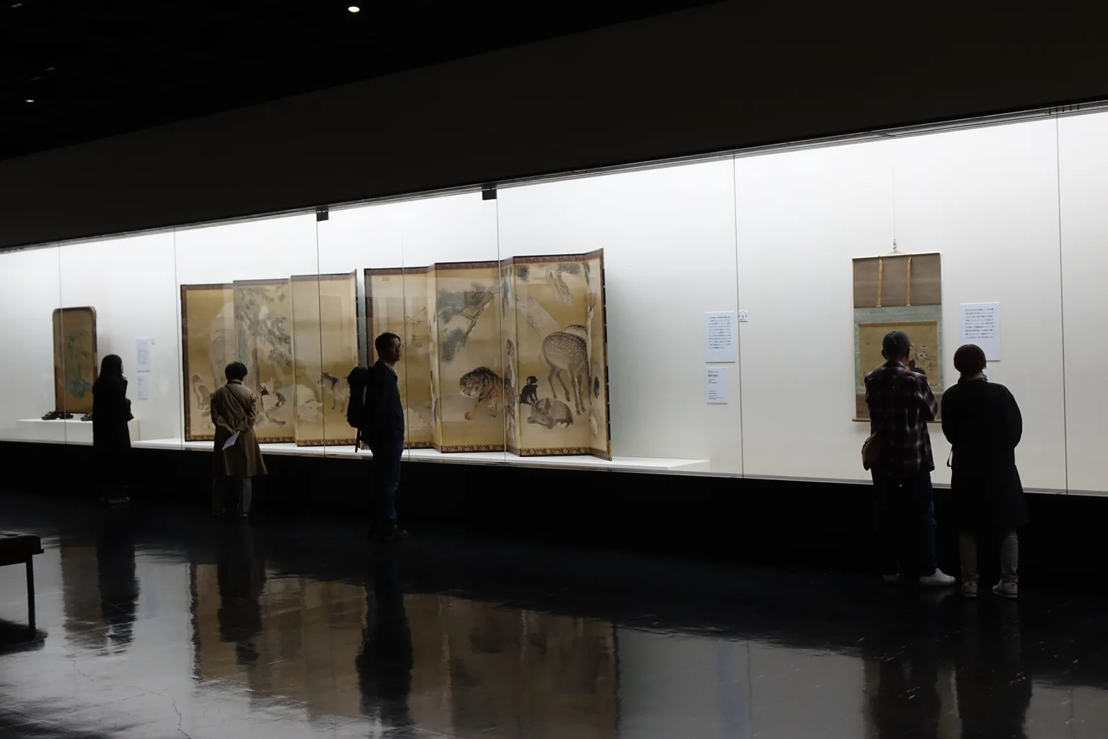 一樓展示室，長向的玻璃展示櫃內陳列屏風與掛軸，深色地面反射出展示櫃的燈光，參觀者沿展示櫃觀覽