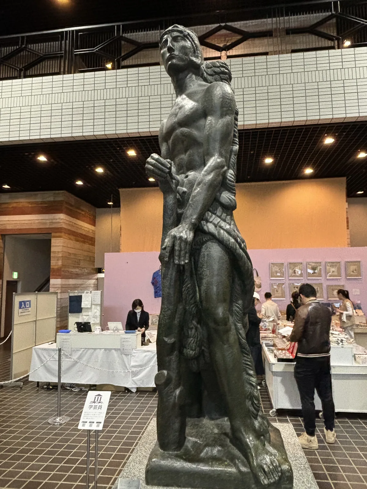 青銅雕像《力》的近景，雕像後方為特別展的商品販售區，白色長桌上陳列商品，背景牆面以多色石材水平條帶砌成