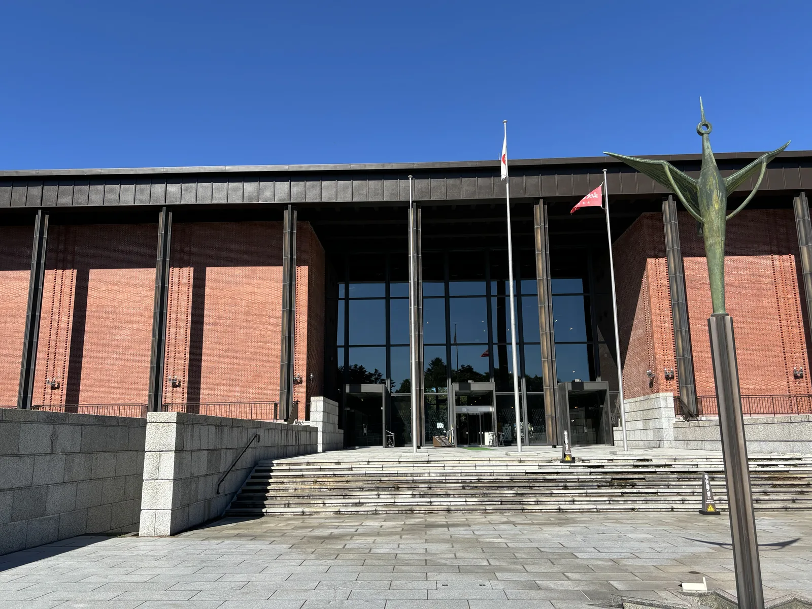 北海道博物館南立面，紅磚方形量體前方為一排深色細柱，中央為玻璃入口，前方為花崗岩擋牆與階梯，右側立著支柱頂端的鶴雕塑，天空湛藍