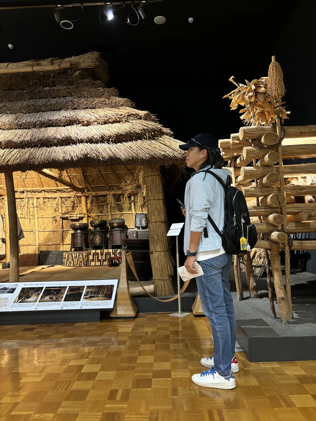 總合展示室內部，深色天花下的聚光燈照亮一座茅草屋頂的復原家屋與一旁的木構倉庫，地面為木質拼花