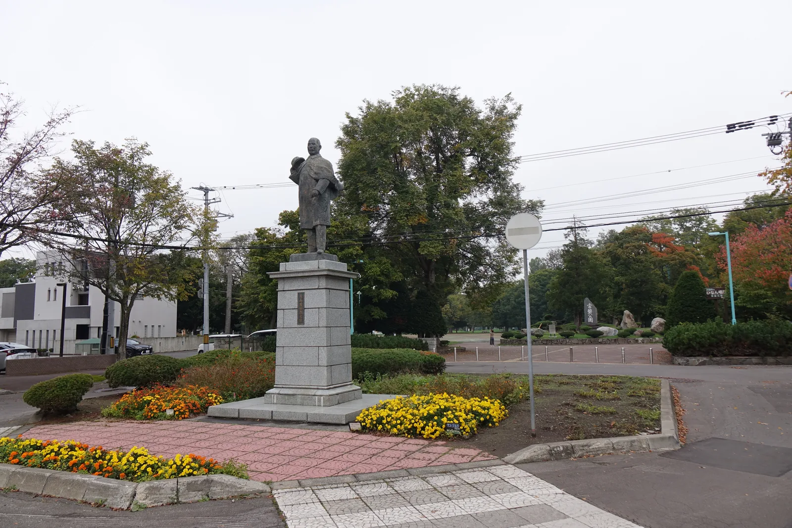 公園入口的圓形花壇，中央為高台座上的永山武四郎青銅立像，四周種植橘色與黃色的花，右後方為公園樹林與石碑