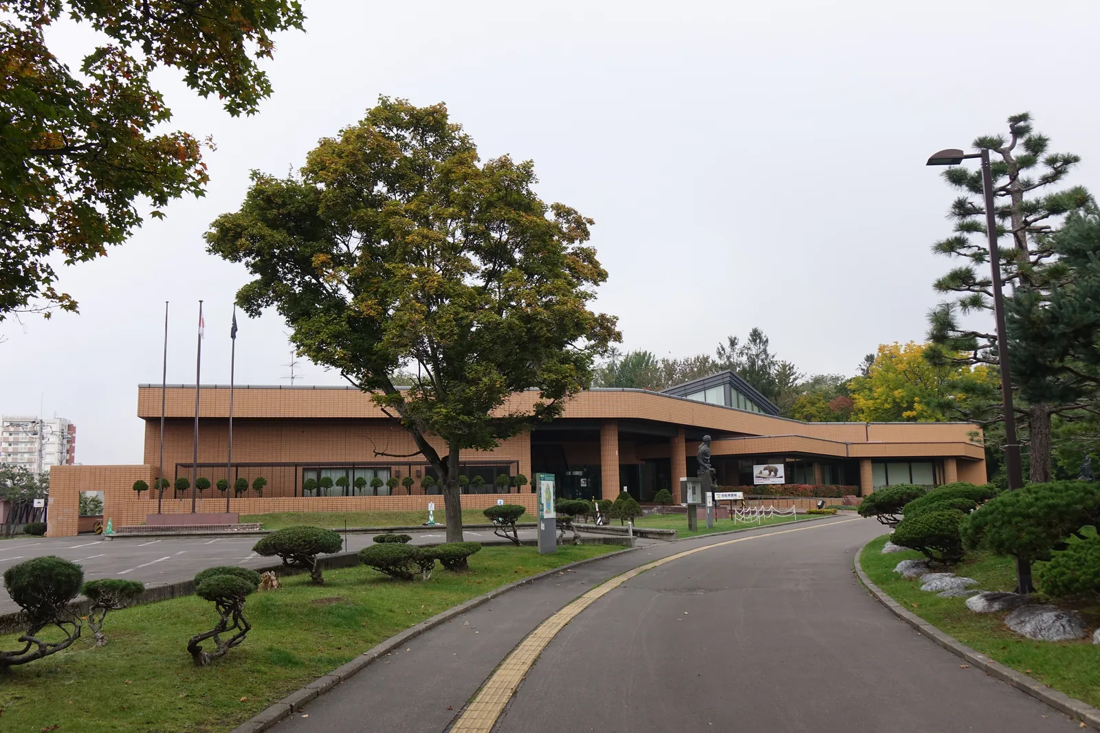 美術館正面與車道，車道以緩弧彎入，兩側為造型松樹，建築外牆為紅褐色磁磚，左側為實牆與旗竿，右側為雨庇下的入口