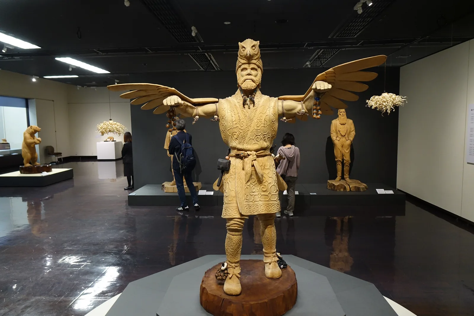第一展示室，深色天花與磨光深色地面，中央八角形台座上立著一尊張開雙翼的大型木雕人像，後方為其他木雕與牆邊展示櫃