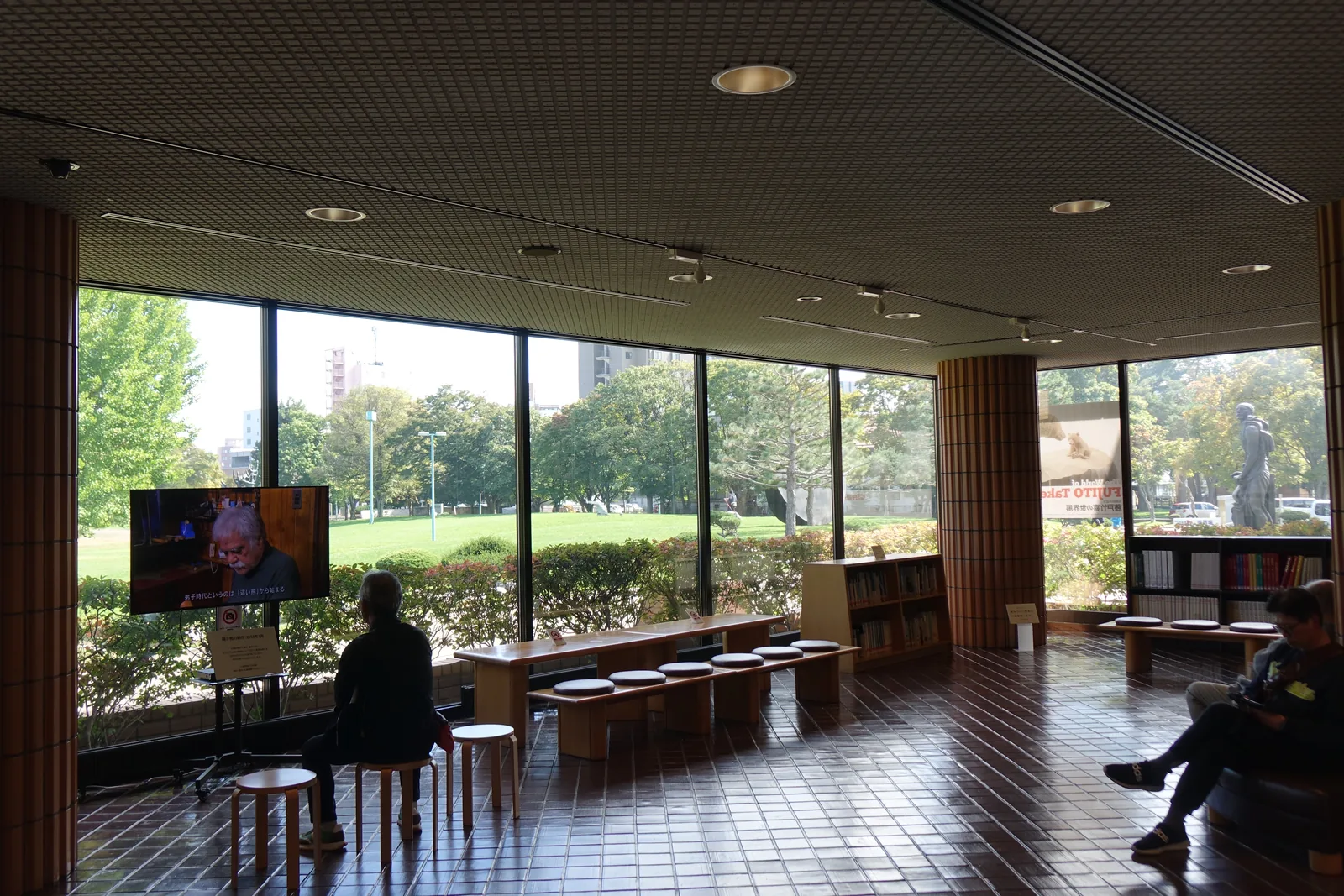 大廳面向公園的休憩區，整面落地玻璃外為草坪與樹林，玻璃前放置木製長凳與圓形坐墊，右側有矮書架與磁磚圓柱，一位訪客坐在凳上觀看電視