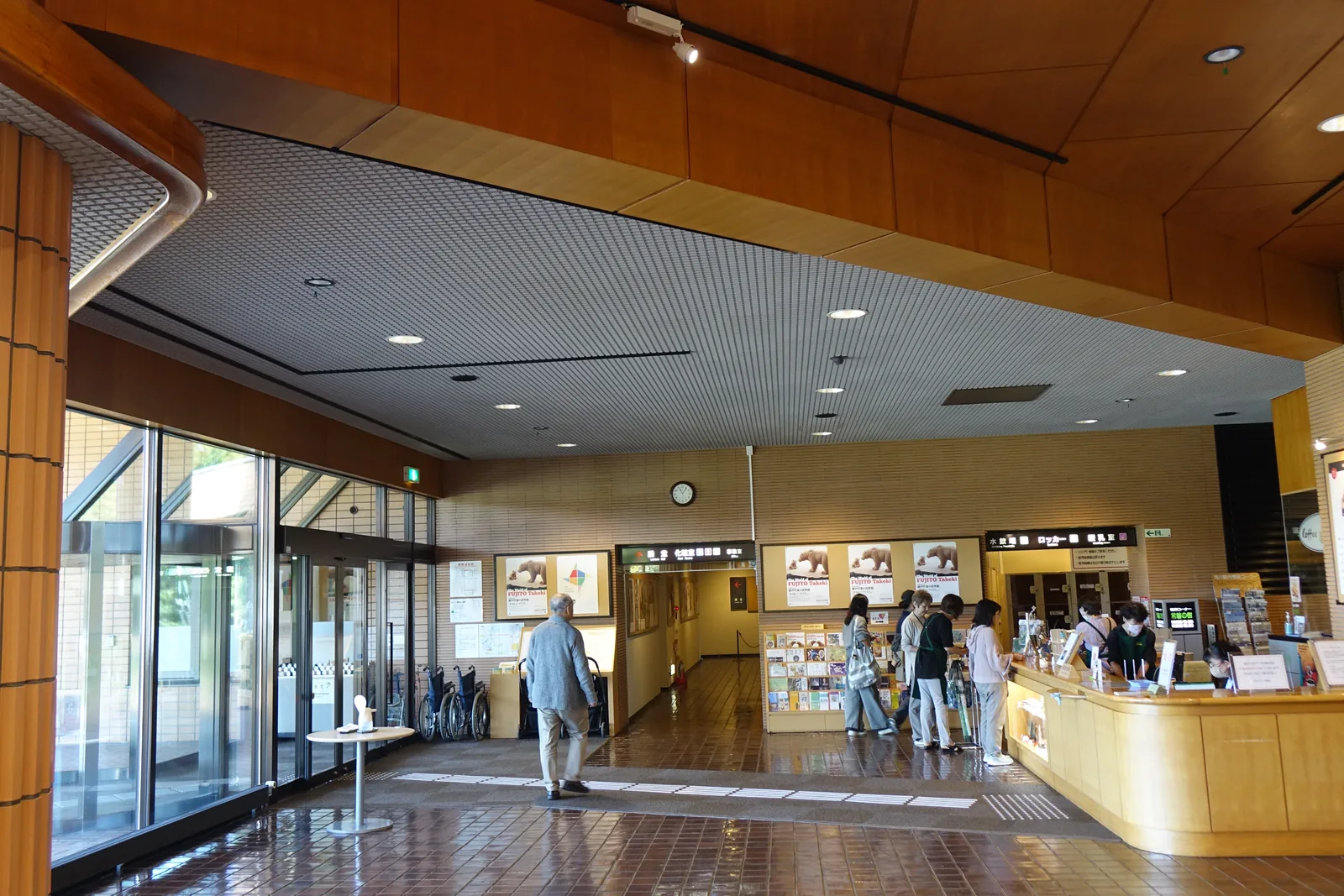 入口大廳，左側為玻璃自動門，中央為服務台與簡介架，訪客在櫃台前詢問，天花分為淺色板材與上方褐色木質面板兩層