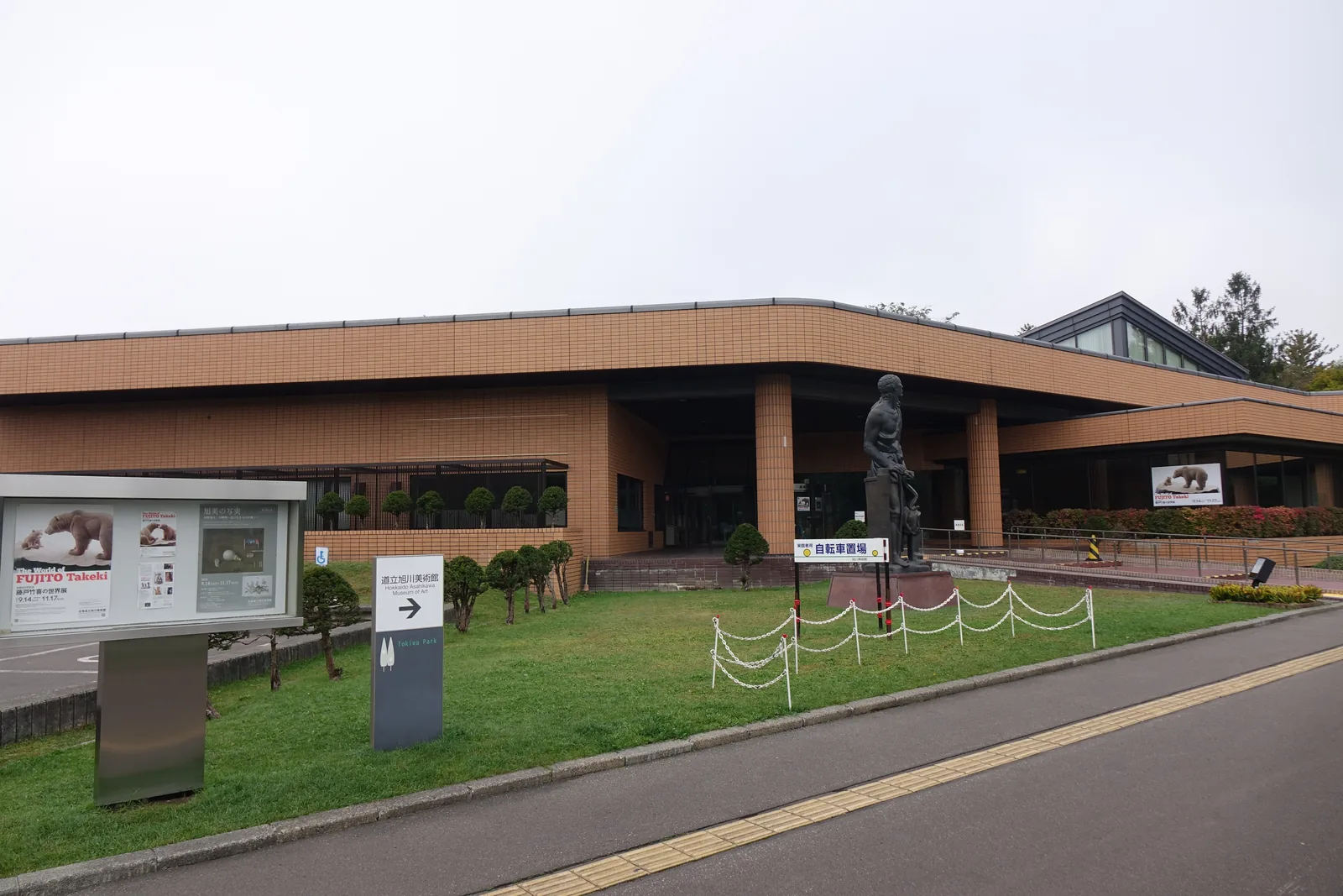 自園路望向美術館，前景為草坪、展覽看板與指示牌，紅褐色磁磚量體的入口雨庇下立著青銅像，右後方為玻璃山形屋頂