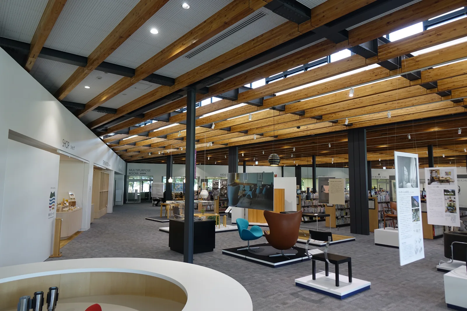 館內全景，前景為圓弧形座檯，中景為家具設計展示區的各式椅子與展示板，左側為商店入口，遠處為多目的室與書架，頭頂為落葉松梁與深灰色鋼梁