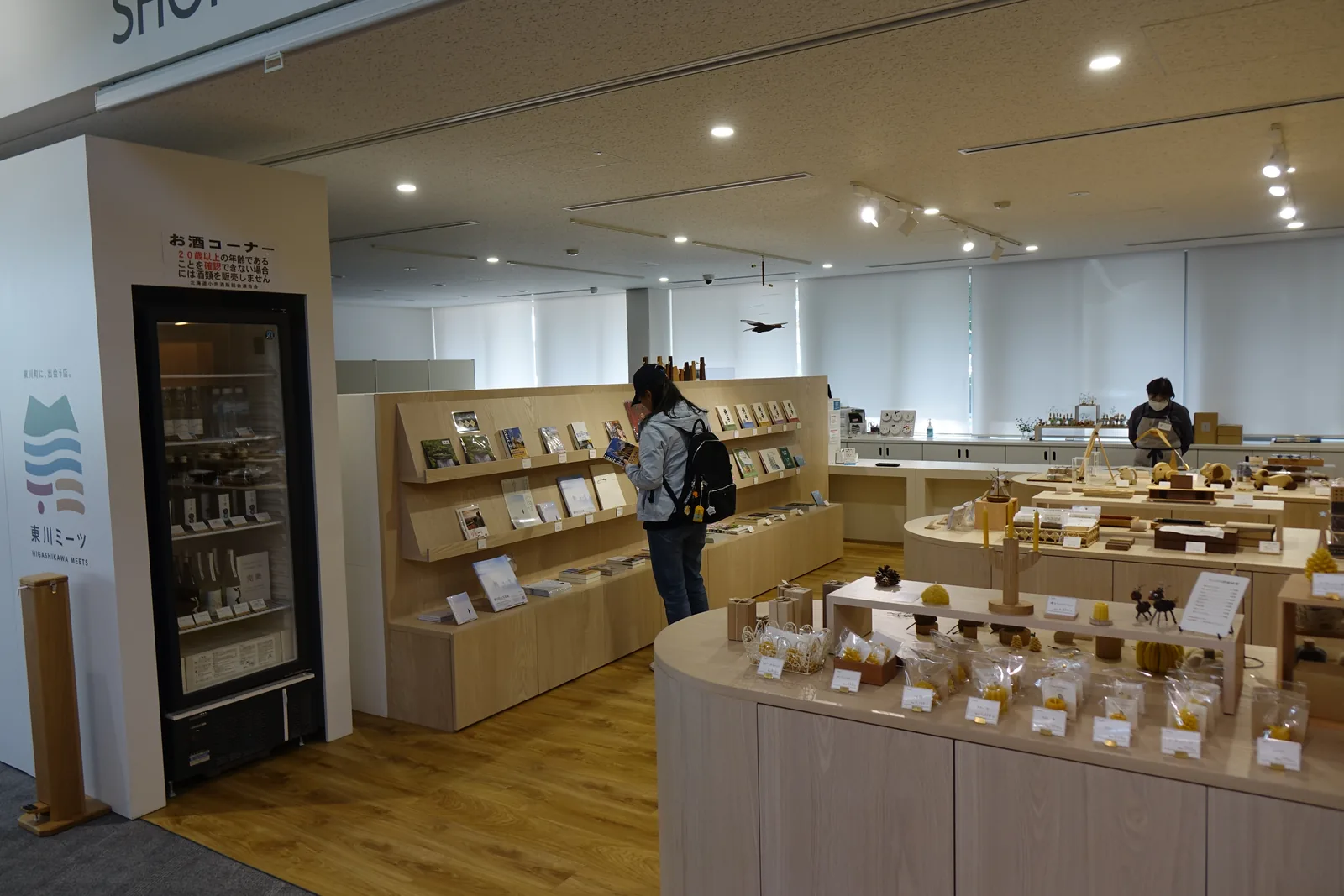 商店內部，淺色木材的弧形展示台上陳列蜜蠟燭與木工藝品，左側牆邊為書架與冷藏櫃，天花為白色平頂