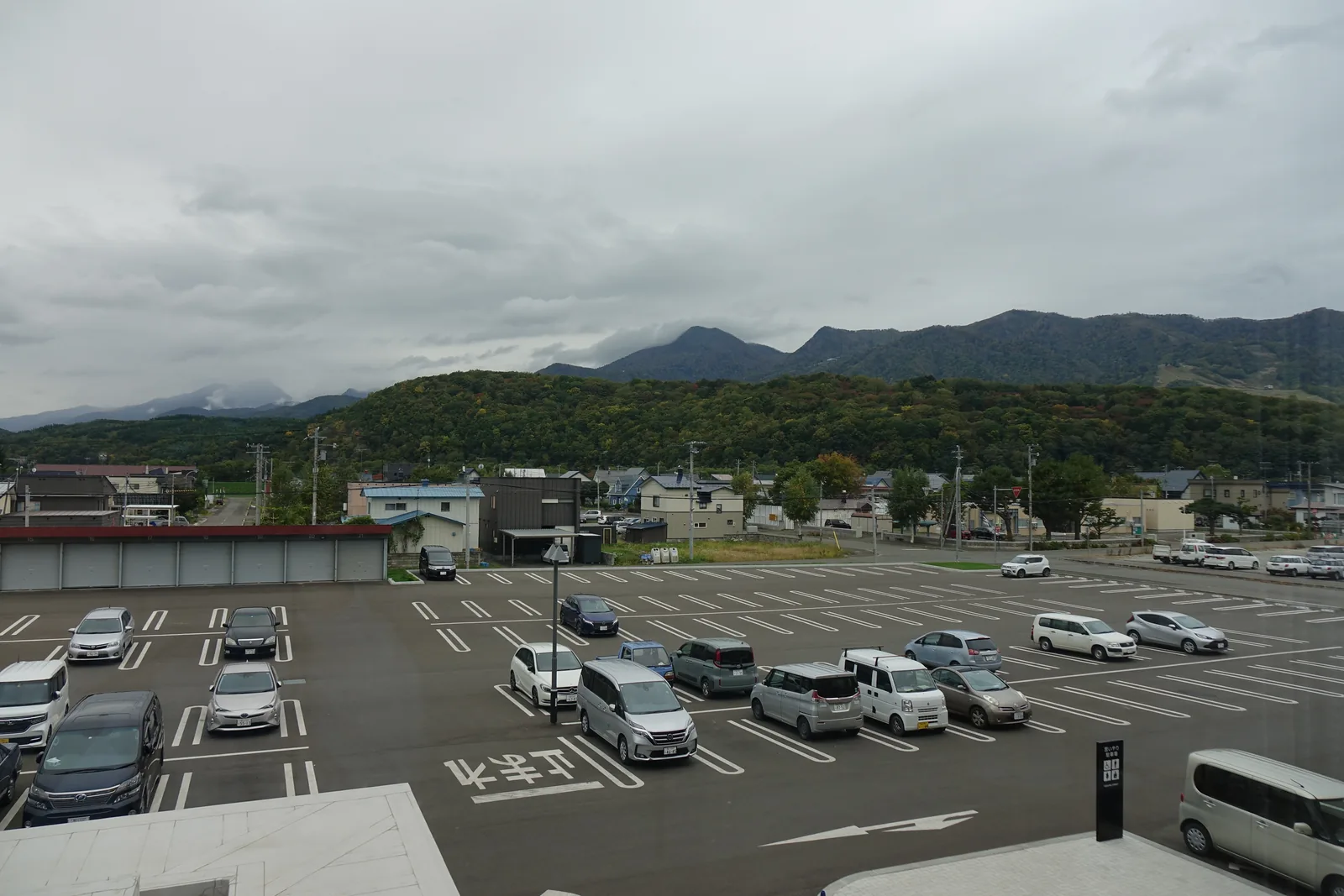 自二樓窗戶望向南側停車場，停車場外為低層住宅屋頂，遠方為環繞盆地的山稜與低垂的雲
