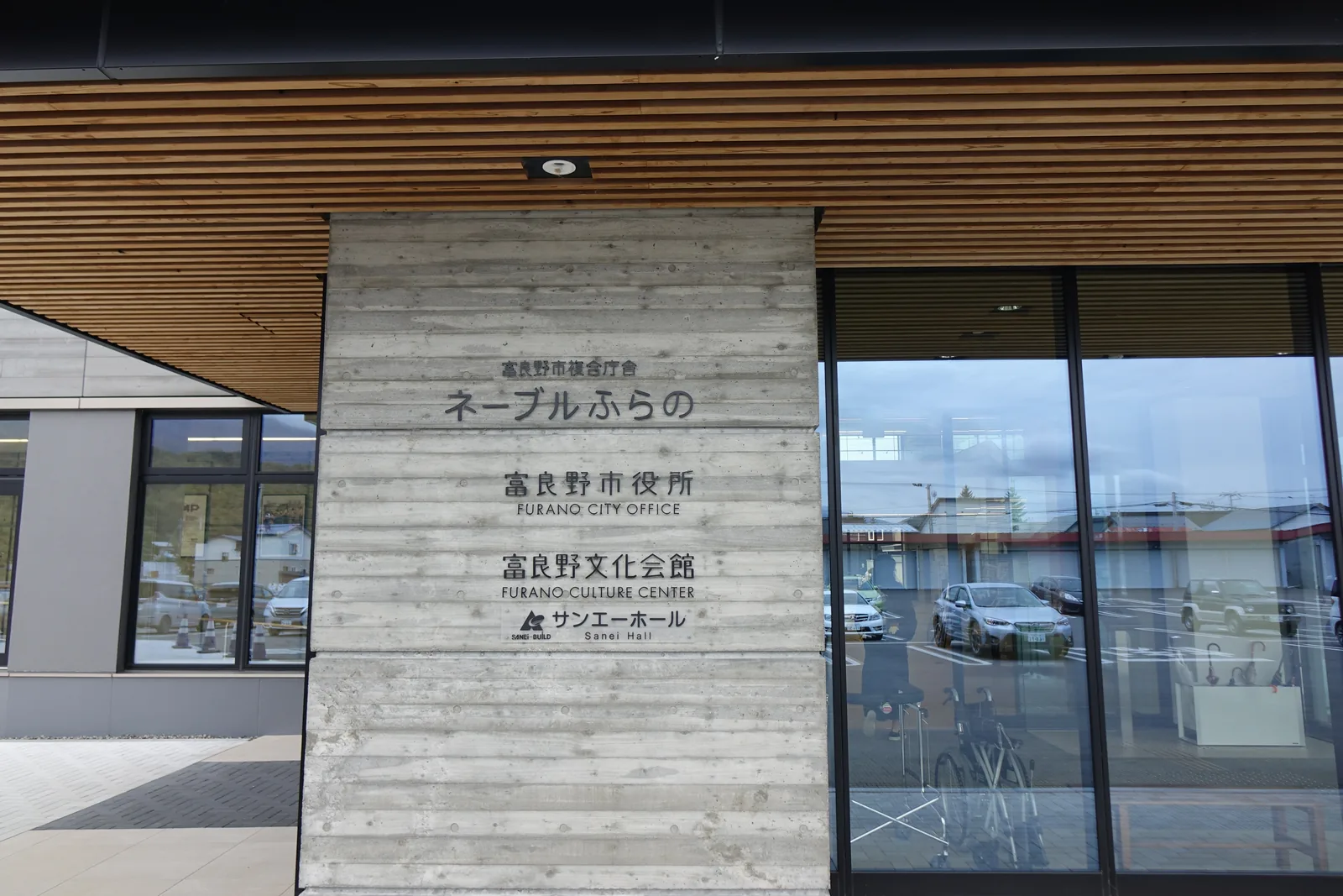 入口雨庇下的館名牆，杉板模板混凝土牆面上寫著建築暱稱、市役所、文化會館與 Sanei Hall，上方為木質條板的雨庇底面，右側為玻璃門