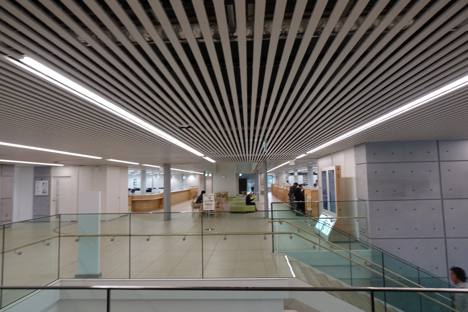 二樓市民大廳，前景為圍住樓梯開口的玻璃扶手，遠處為窗口櫃台與等候座位，右側為模板混凝土牆，頭頂為白色條板天花與線性照明