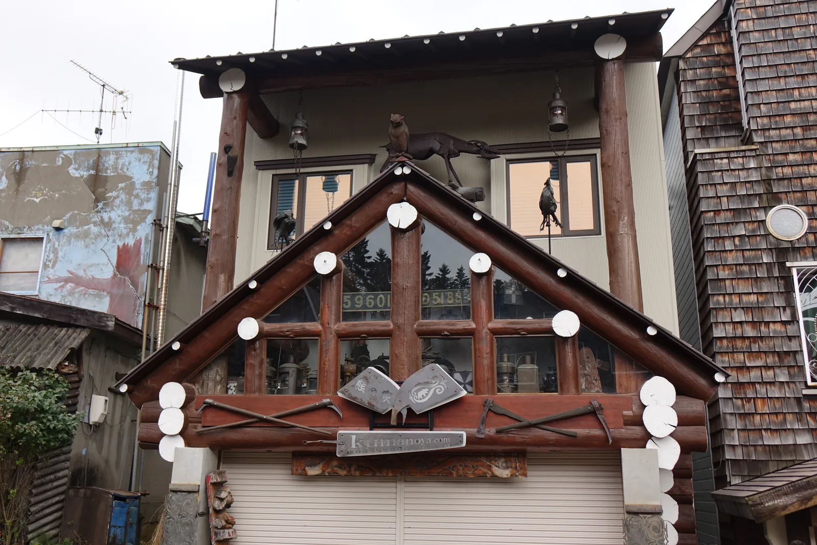 愛努聚落內的一棟民藝店，二層建築的正面以原木組成山牆與窗框，山牆上嵌著白色圓盤與金屬雕刻，屋簷下掛著鈴與動物剪影