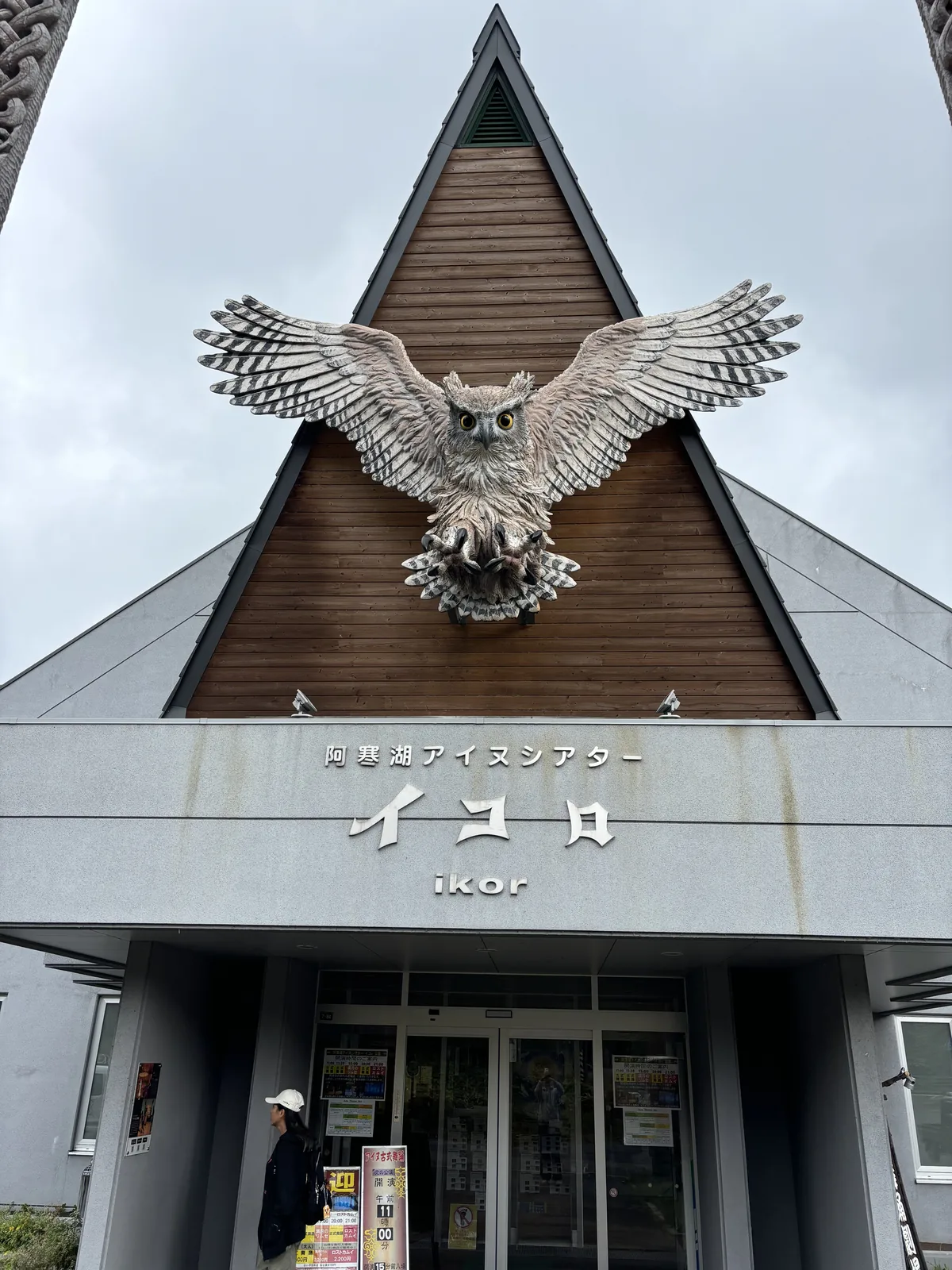 劇場正面入口，上部為三角形木板山牆與貓頭鷹木雕，下部為灰色量體與館名，入口退在平屋頂雨庇下，兩側立著雕刻柱