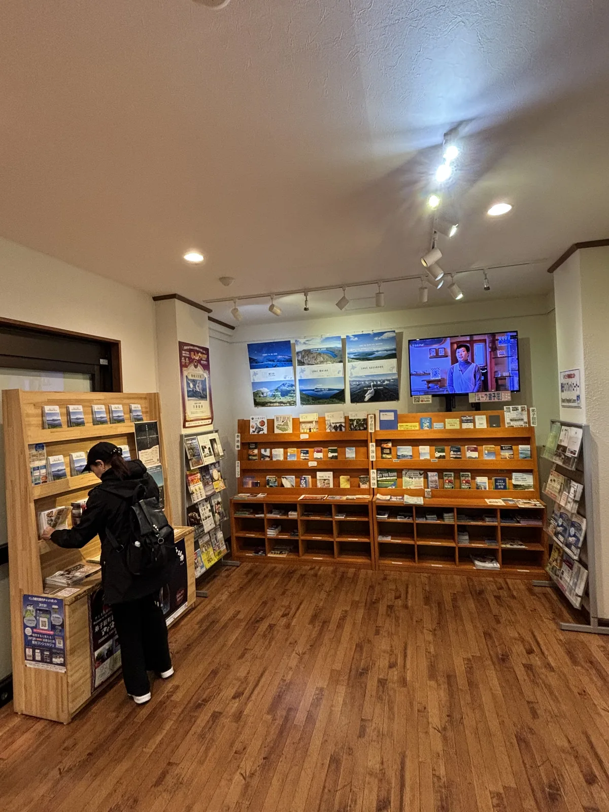 觀光案內所，木製書架沿牆排列陳列導覽手冊，牆上掛著摩周湖的照片與電視螢幕，天花設有軌道燈，地面為木地板