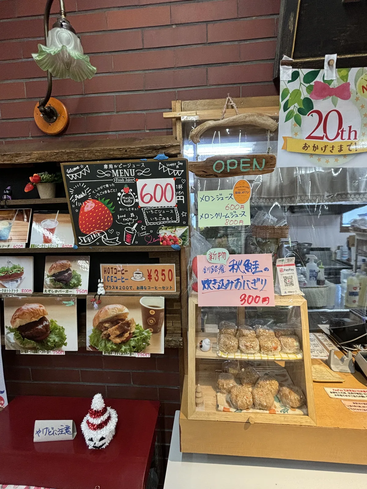 以紅磚牆為背景的take-out櫃台，牆上掛著壁燈與手寫菜單板，櫃台上陳列漢堡照片與飯糰，右側玻璃櫃內擺放點心
