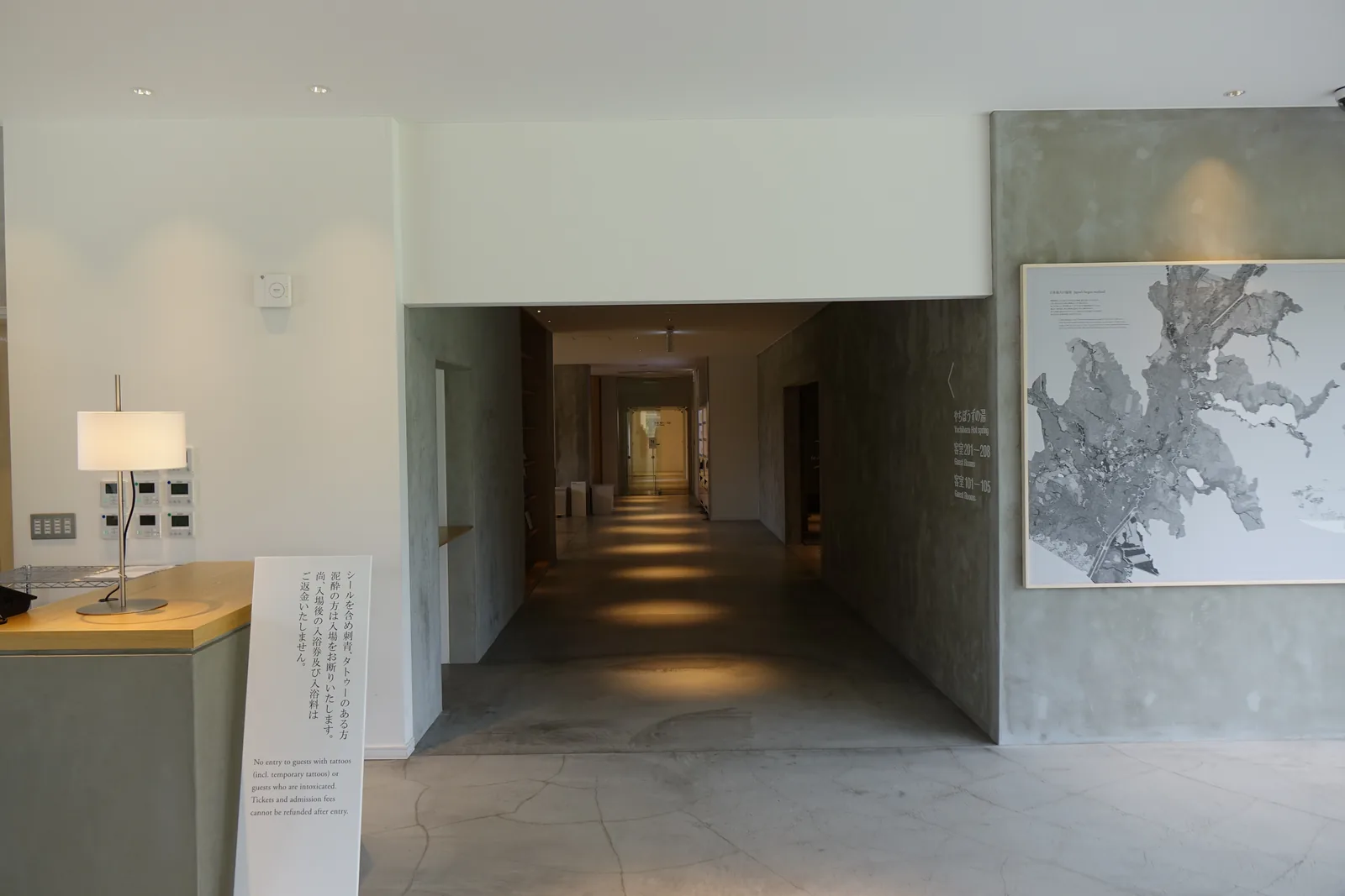 走廊開口的正面，灰色牆面圍出的長廊向深處延伸，地面以間接照明打出光斑，右側牆上為黑白的濕原地圖