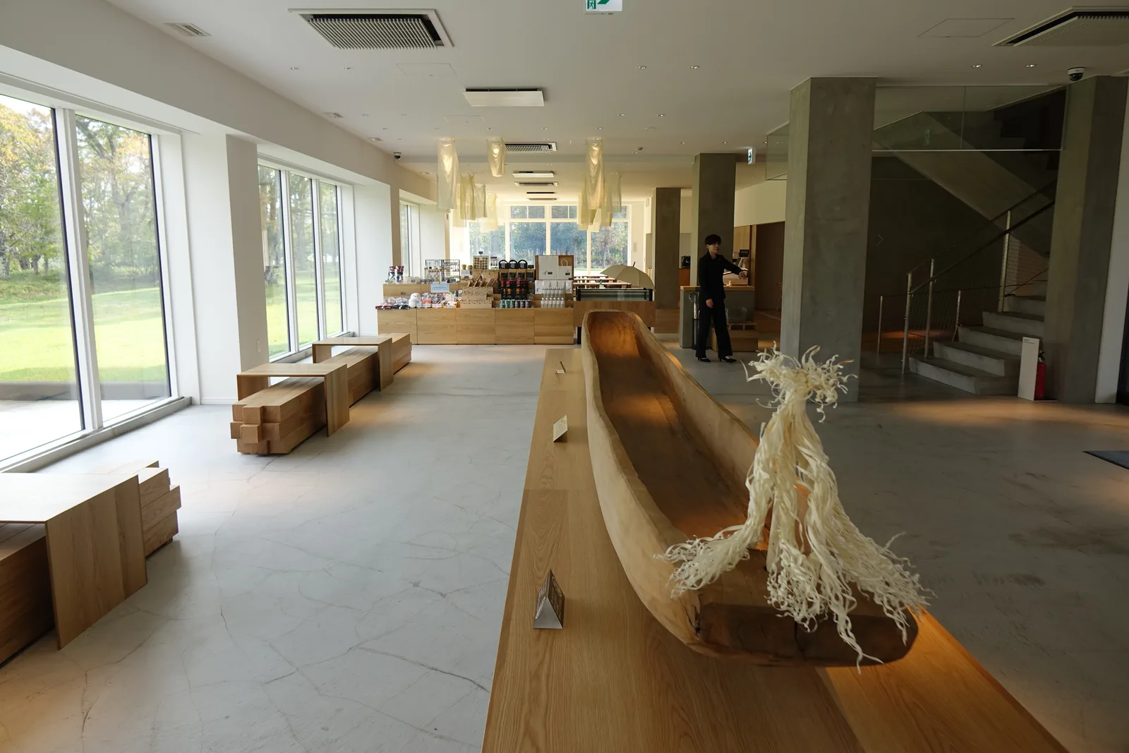 大廳全景，中央為長條木製台座上的獨木舟與白色纖維造形，左側為落地窗與木製座椅，深處為商店與懸掛的布燈，右側為混凝土柱與樓梯
