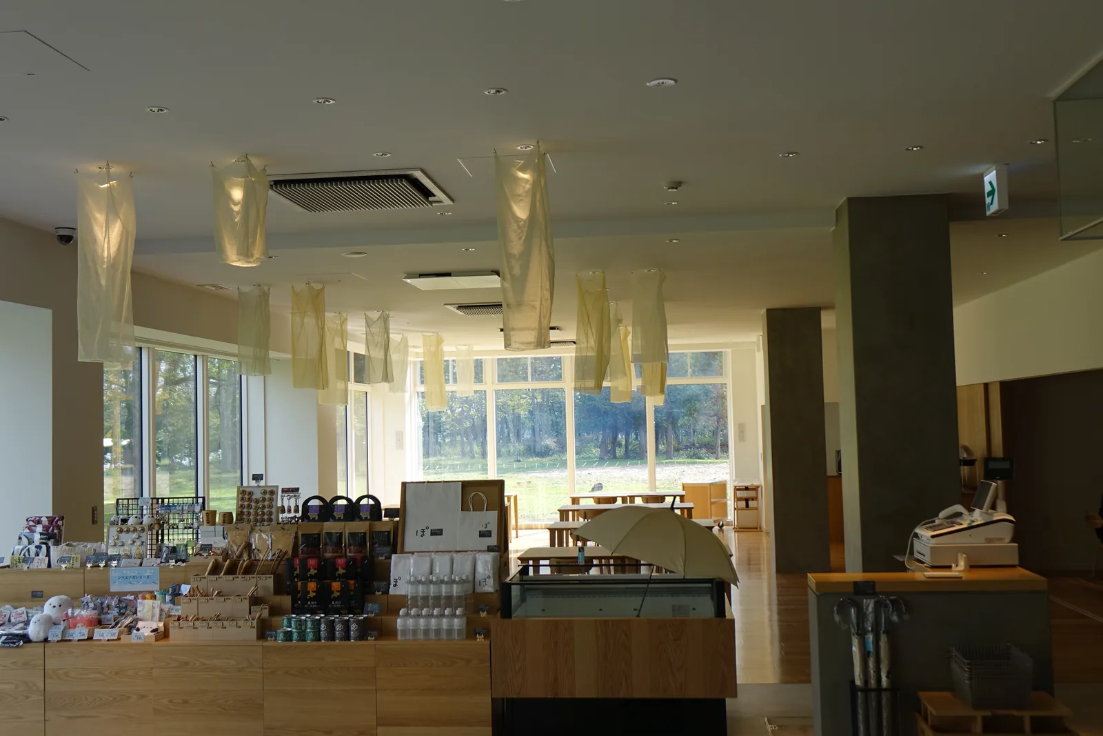 商店，木製展示台上陳列食品與雜貨，天花下懸掛數個半透明布料的長形燈具，深處為落地窗與餐廳的桌椅