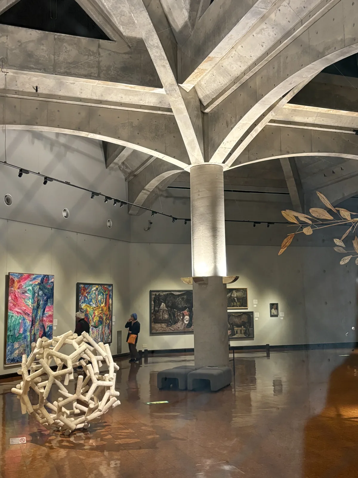 展示室內部，中央的混凝土圓柱向四方分出彎曲的拱肋，與相鄰柱子的拱肋在天花交織，牆面掛著油畫，地面反射燈光，前景為白色的木條球形作品