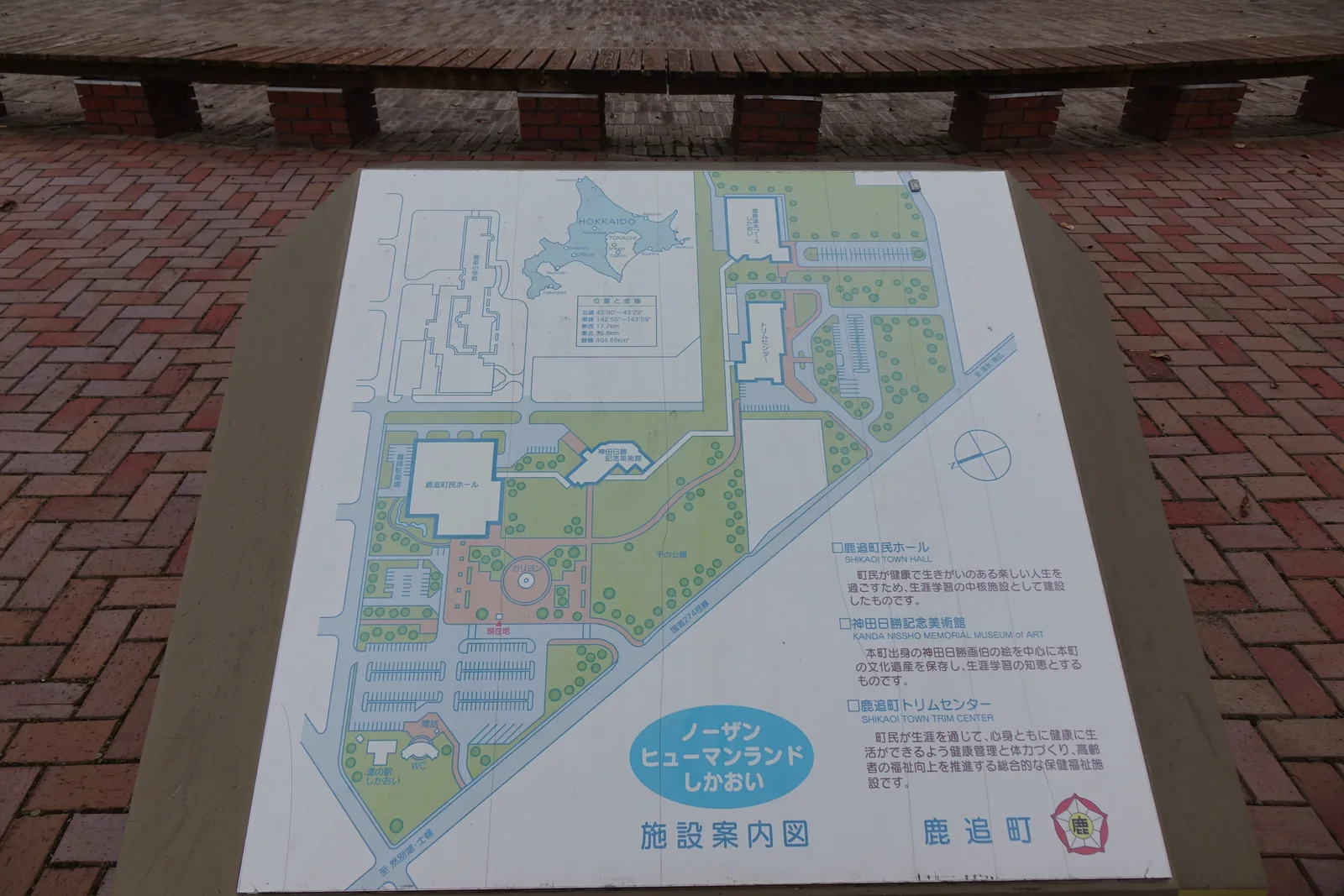 現場的設施案內圖，以平面圖標示町民會館、神田日勝記念美術館、Trim Center 與公路休息站的位置，右下為各設施的說明文字，前方為紅磚鋪面與木製長椅