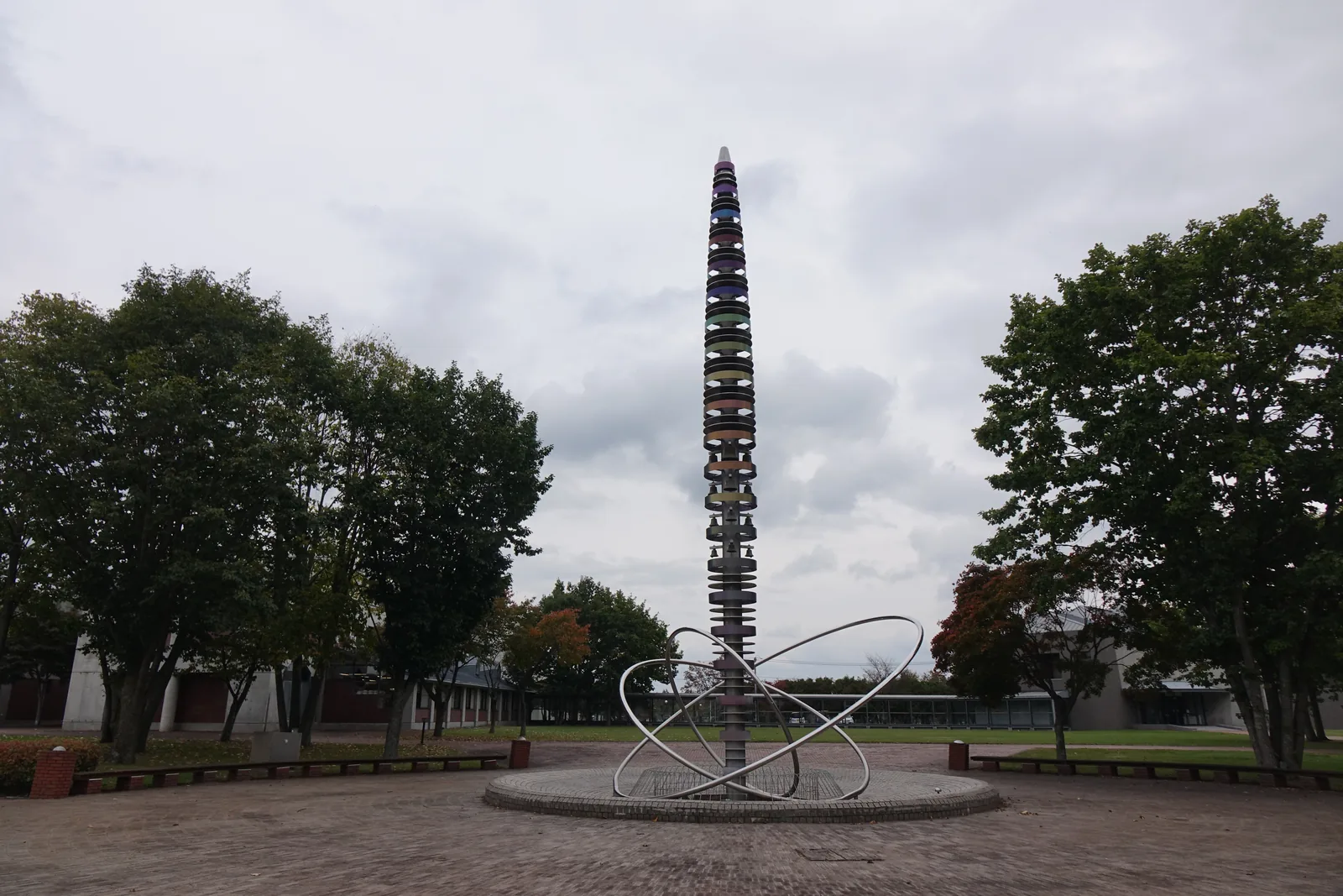 紅磚鋪面的圓形廣場，中央立著一座多層的 carillon 塔，底部有金屬環形雕塑，周邊為樹木與遠方低矮的建築