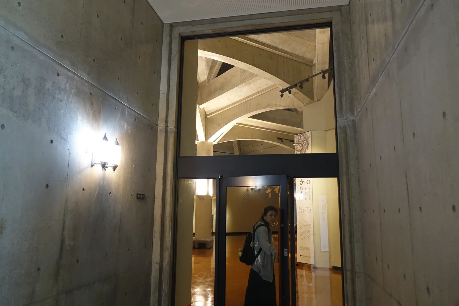 展示室入口，兩側為清水混凝土牆與壁燈，入口上方的玻璃透出展示室天花的混凝土拱肋，下方為玻璃門