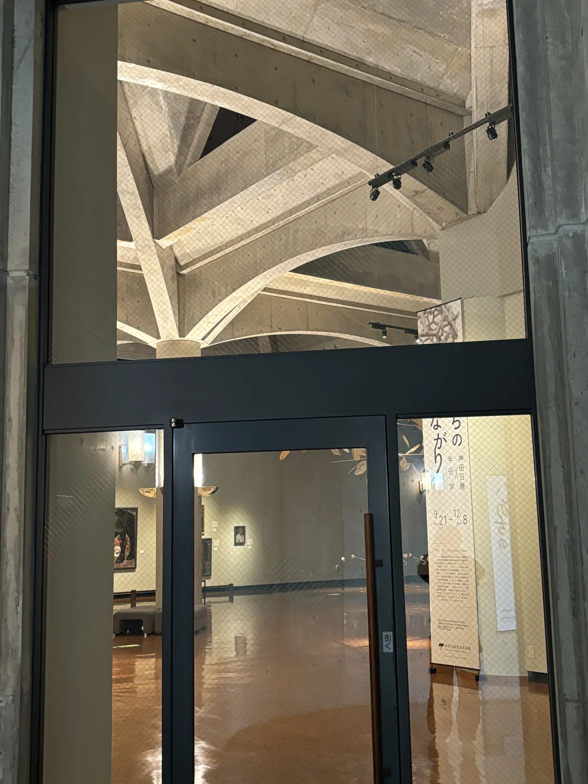 展示室入口的玻璃門，上方可見彎曲的混凝土拱肋與軌道燈，門內為光亮的地面與展示牆，右側為展覽海報
