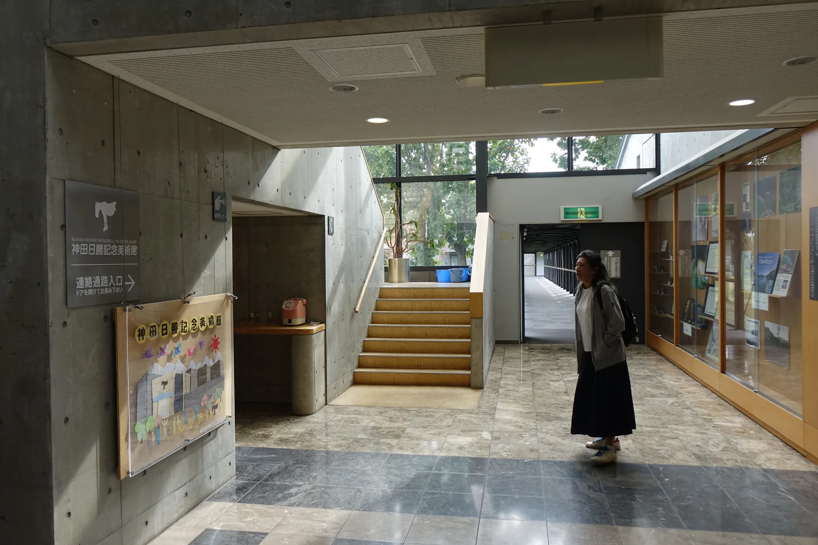 連絡通路入口，左側清水混凝土牆上掛著館名標示與連絡通路入口的指示，正面為上行的樓梯，右側為玻璃展示櫃與通往町民會館的長廊