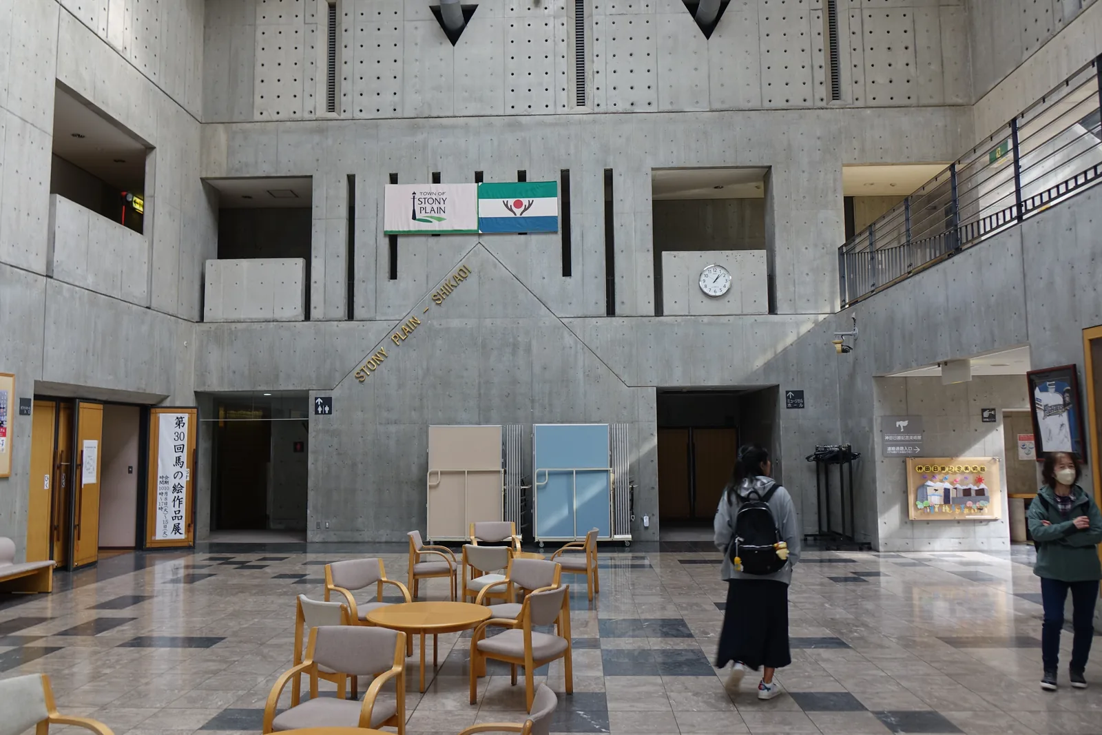 町民會館的挑空大廳，四周為清水混凝土牆，上部牆面排列模板孔，正面掛著 Stony Plain 的旗幟，二樓的開口與走廊面向大廳，地面放著桌椅，左側立著馬之畫作品展的看板