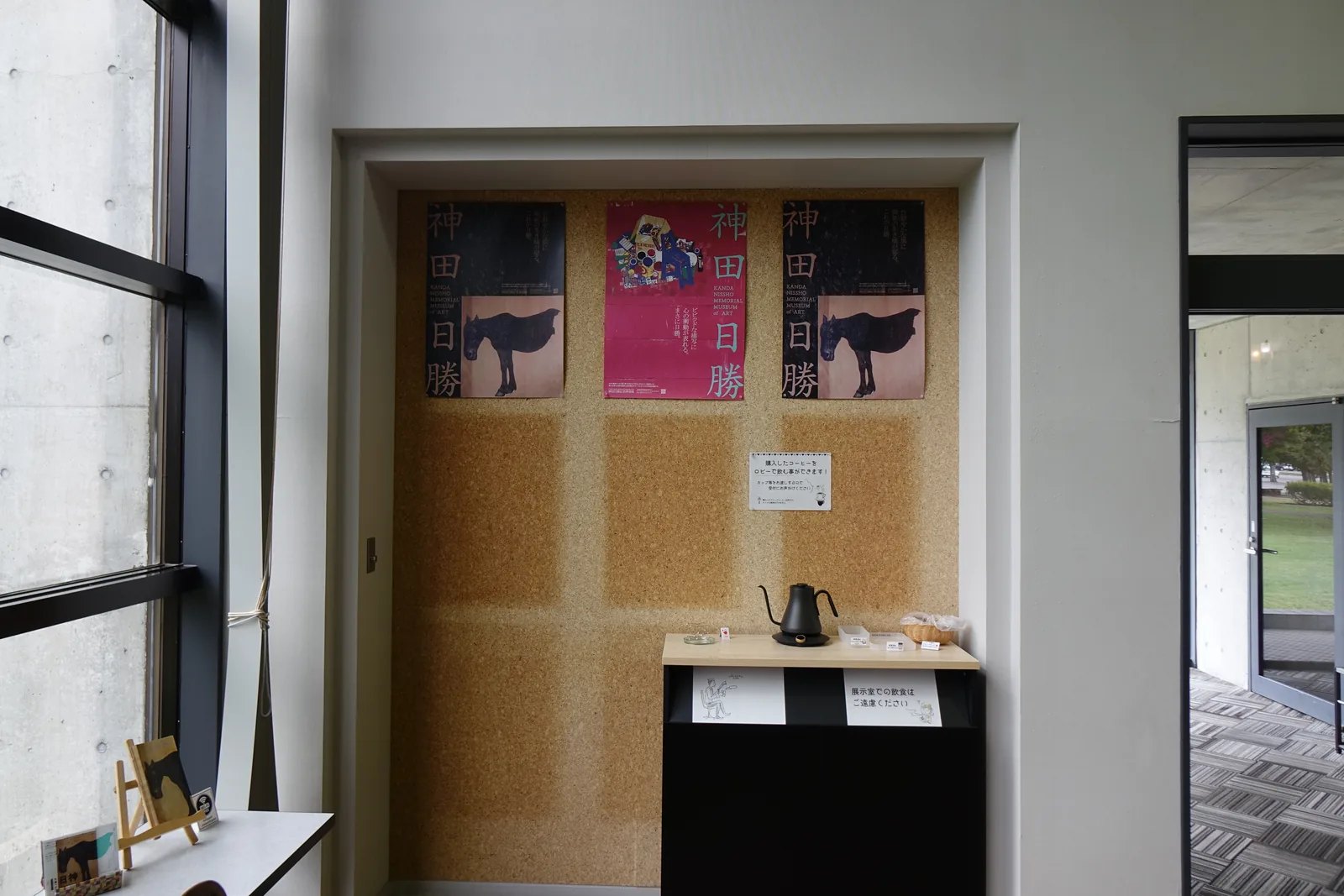 大廳一角的咖啡角，軟木板壁龕上貼著三張神田日勝的展覽海報，下方的黑色小台上放著熱水壺與杯具，左側為大廳的窗