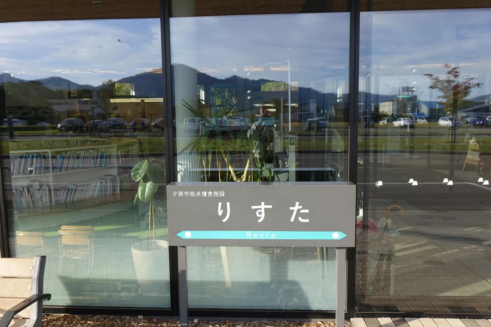 入口旁的館名牌，灰色底板上寫著館名，下方為藍綠色的箭頭帶與英文 Resta，背後的玻璃映出遠方的山與停車場
