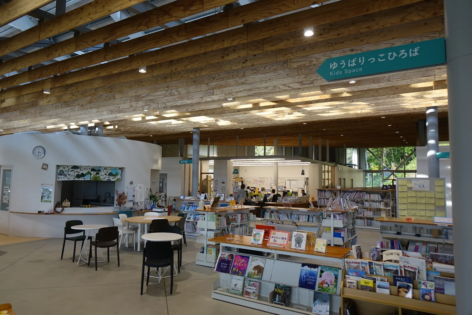 Kids Space，左側為白色圓弧形量體與附水槽的櫃台，中央為圓桌與座椅，右側為繪本架與書架，上方懸掛綠色的 Kids Space 指標
