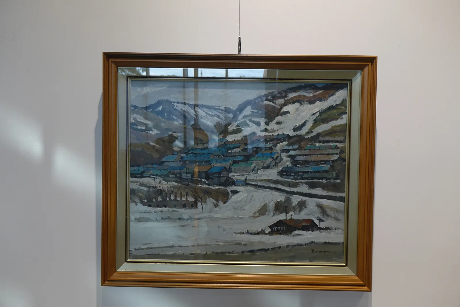畫框中的油畫，描繪積雪山谷中的礦山聚落與藍色屋頂的建築群，背景為白牆