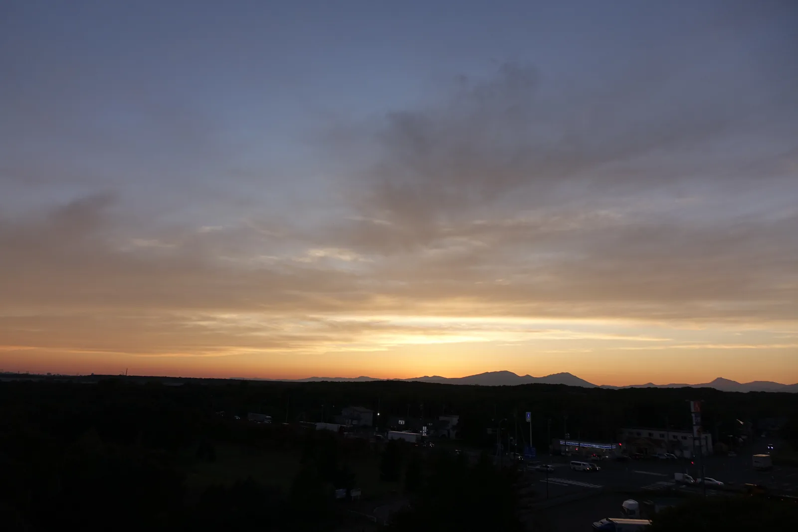 自展望設施屋頂望向西方的夕陽，太陽落在遠方的山稜之後，前景為深色的樹林與市街的建築