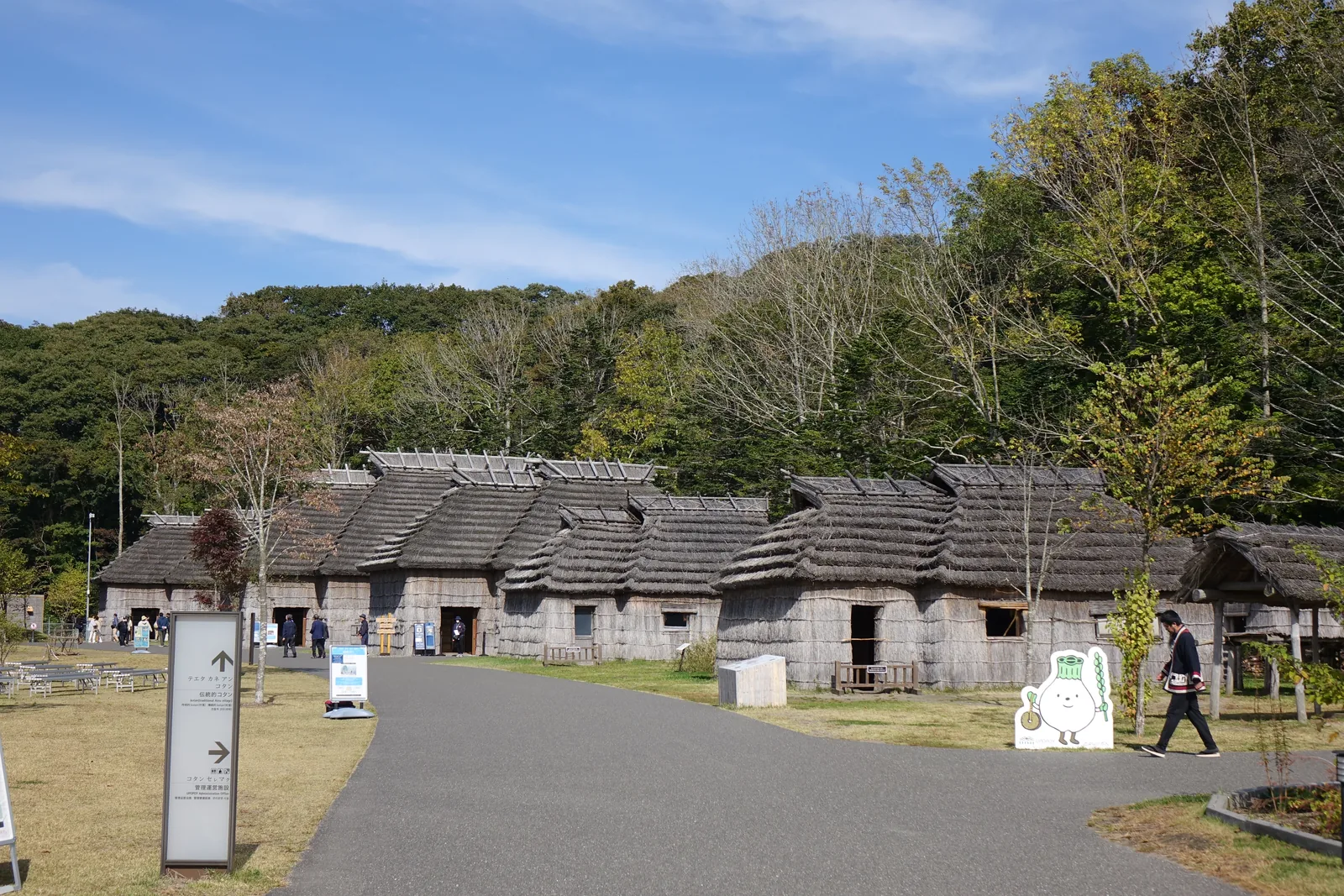 伝統 kotan 的 chise 群，數棟茅草屋頂的家屋沿步道排列，背後為覆滿樹林的丘陵，前方為草地與長椅
