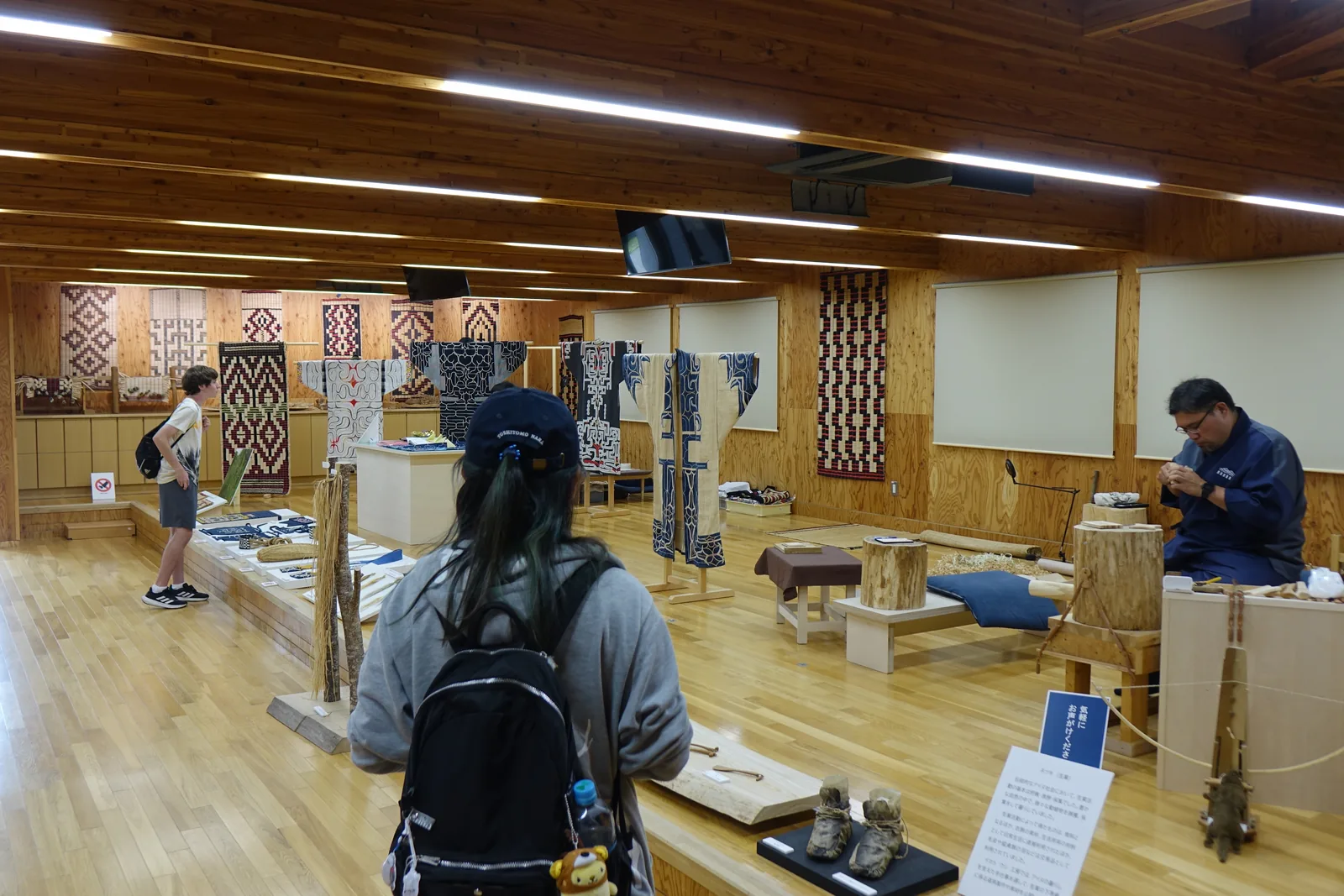 工房內部，木地板上陳列著樹皮纖維、編籃與木製工具，深處為工藝家的作業台與掛在牆上的傳統服飾