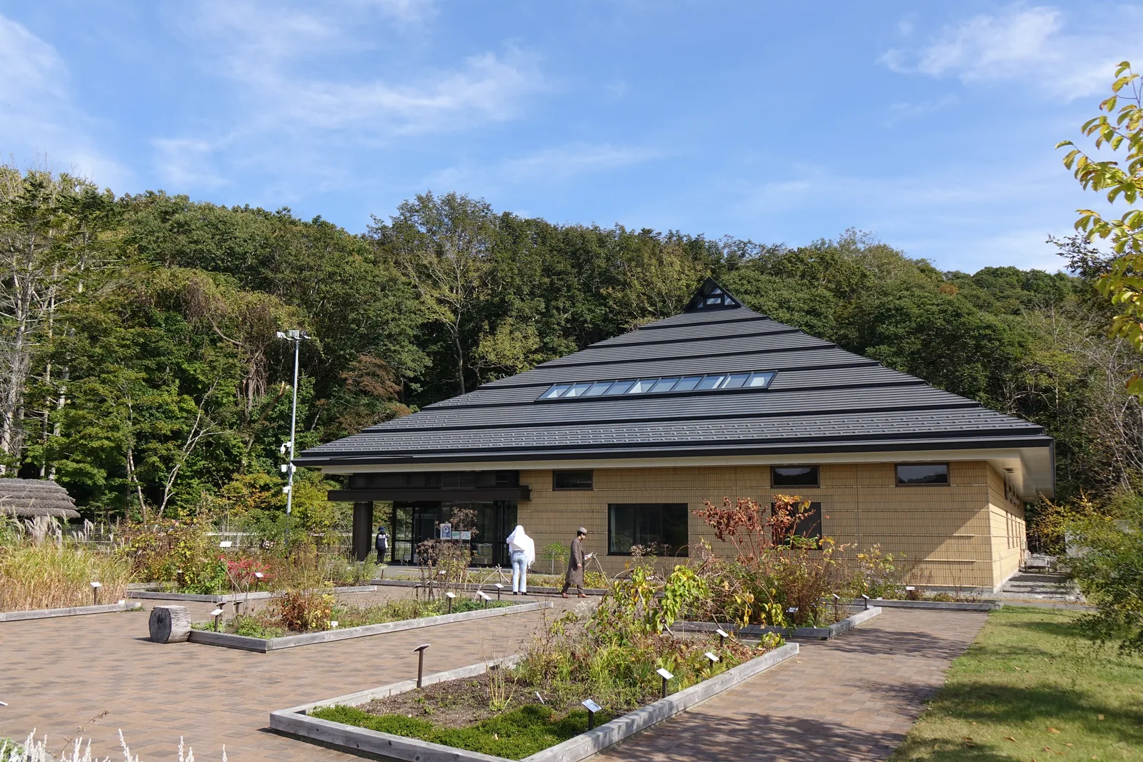 四角錐屋頂的建築，深灰色的屋頂頂端開有天窗，外牆為淺褐色的面材，前方為花圃與磚鋪步道，背後為樹林