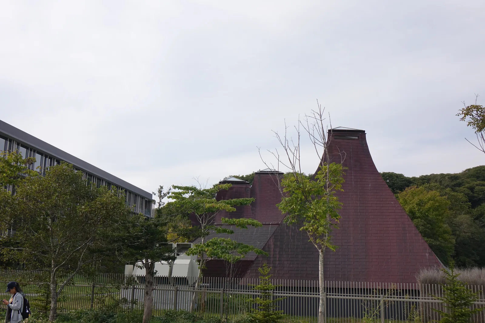 隔著草地與樹木望向界 Poroto，褐色的圓錐形屋頂立在湖畔，左側為灰色的旅館棟，背後為樹林
