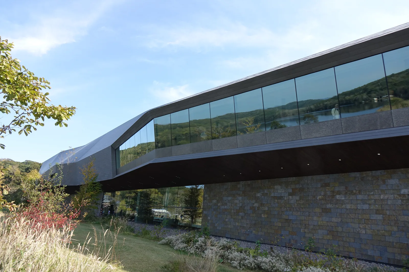 博物館北側立面，黑灰色的長條量體沿湖岸展開，二樓局部為帶狀玻璃，前方為草地、步道與植栽