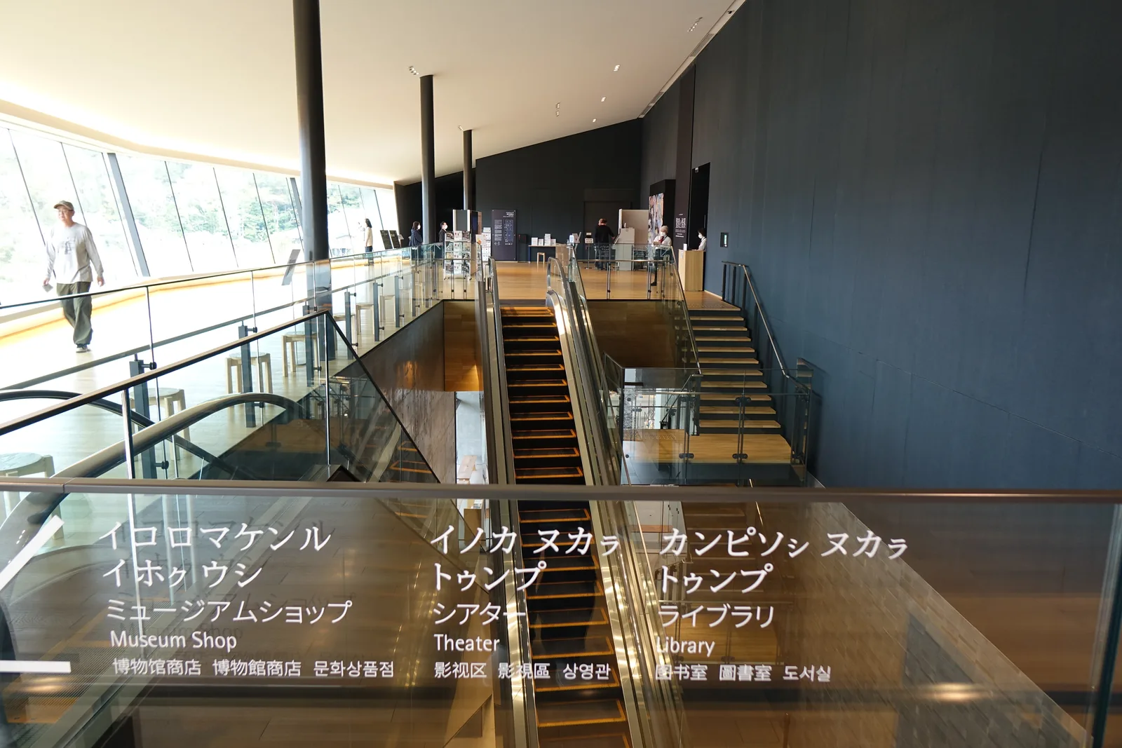 自 Panoramic Lobby 回望挑空，下方為手扶梯與玻璃欄杆的樓梯，右側為深色牆面與細柱，左側為傾斜玻璃