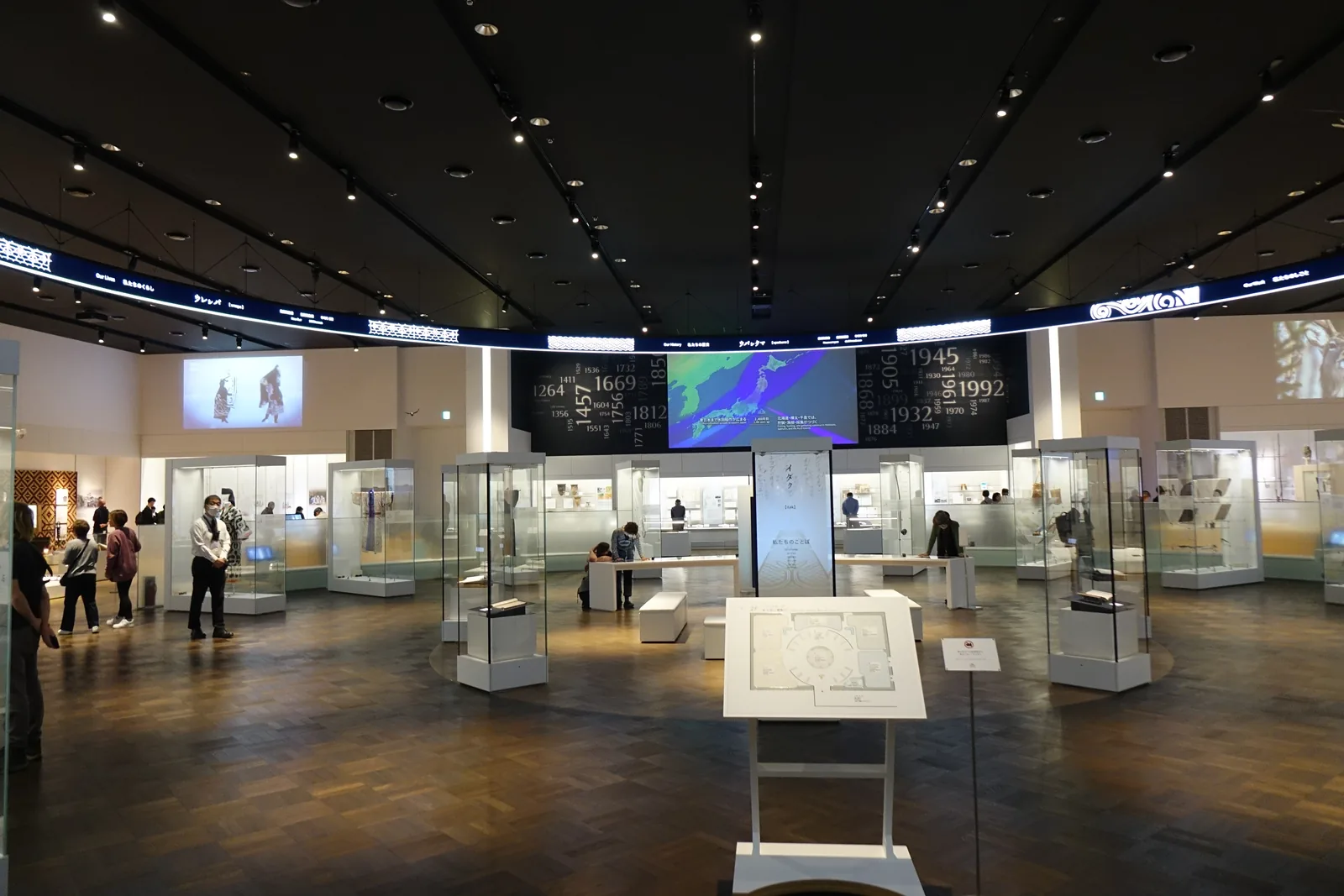 基本展示室，黑色天花下懸吊著環形的光帶與螢幕，中央為環形的展示廣場與低台，四周為玻璃展示櫃，地面為木地板