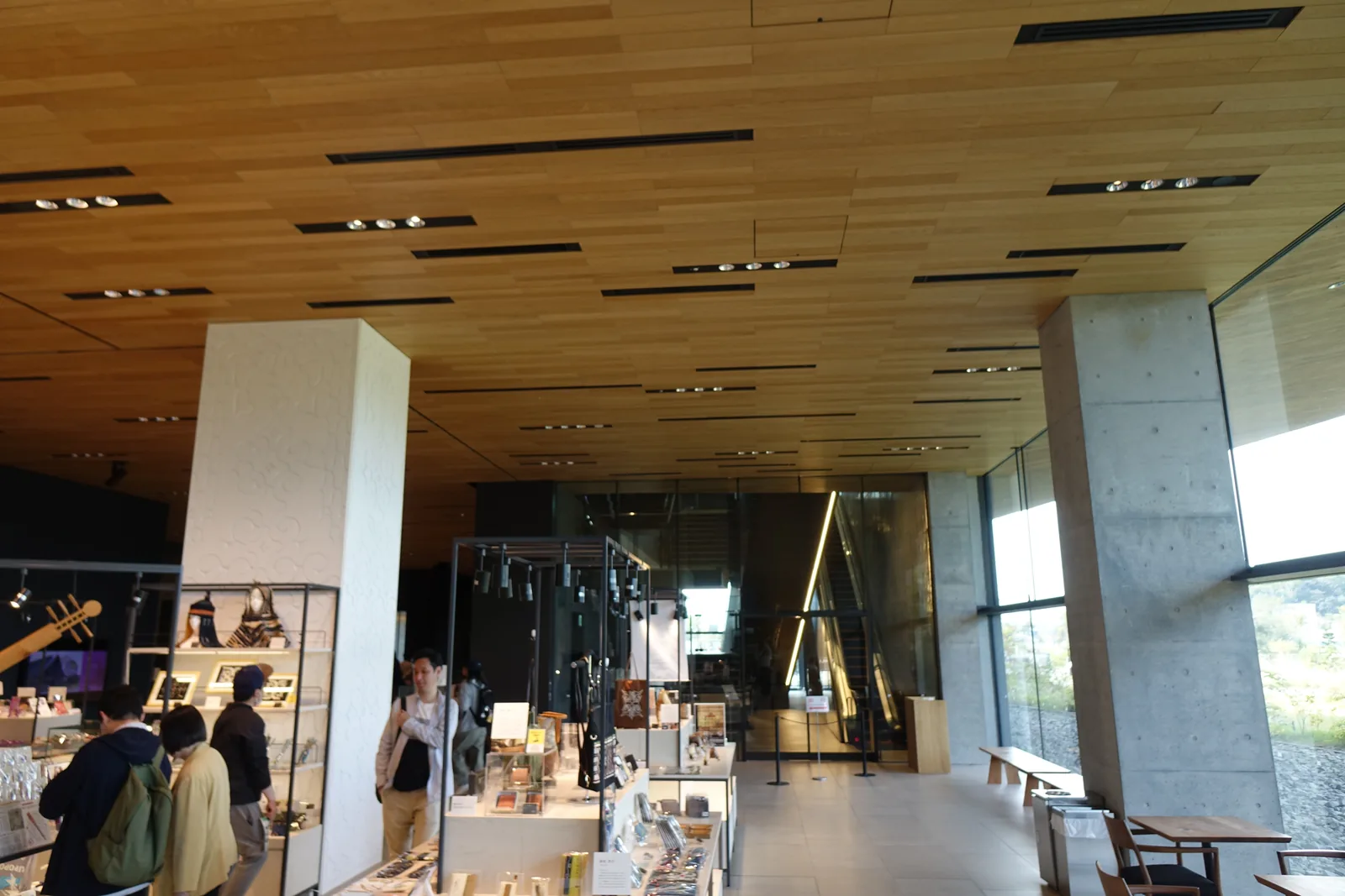 博物館商店，木質天花下為玻璃展示櫃與陳列台，右側為清水混凝土柱與落地玻璃，窗外為草地