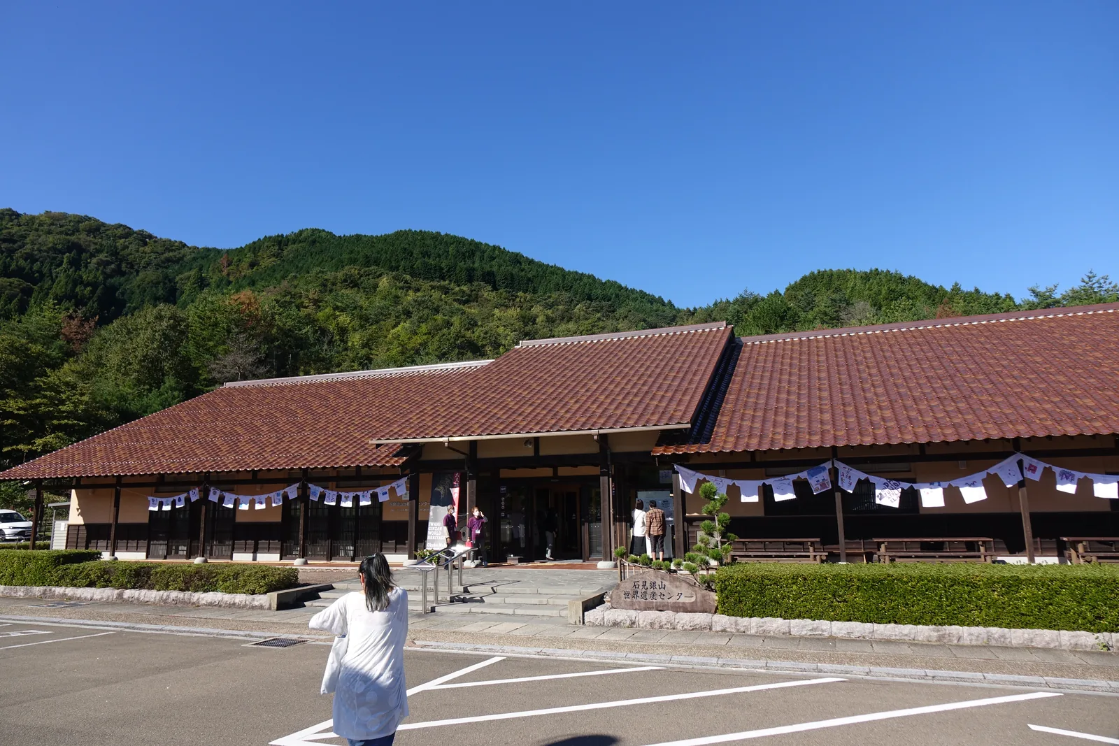 石見銀山世界遺產中心的 guidance 棟正面，赤褐色石州瓦的切妻屋頂連成一長列，中央入口上方另起一道小山牆，簷下是白色土壁與深色木板腰壁，前方是停車場與石砌的花台，背後是覆蓋樹林的山