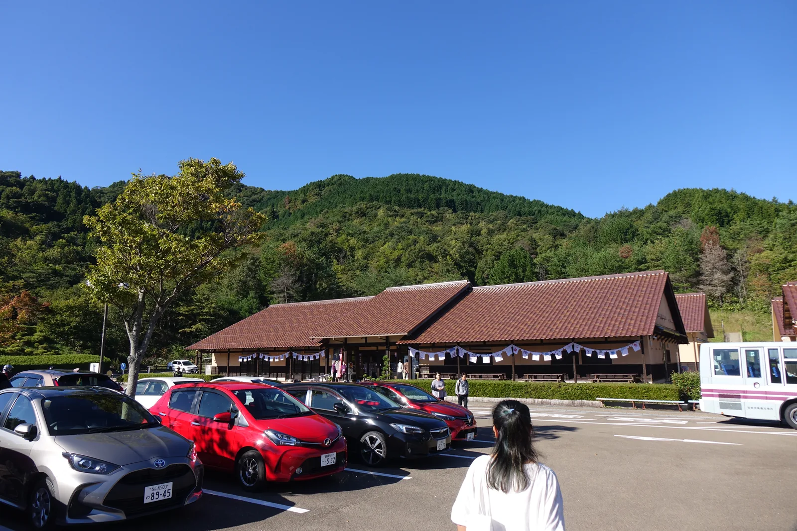 自停車場望向中心，前景是停放的車輛與一輛白色的巡迴巴士，中景是兩道相連的赤褐色瓦頂建築，簷下掛著布幔，背後是樹林覆蓋的山坡與晴空