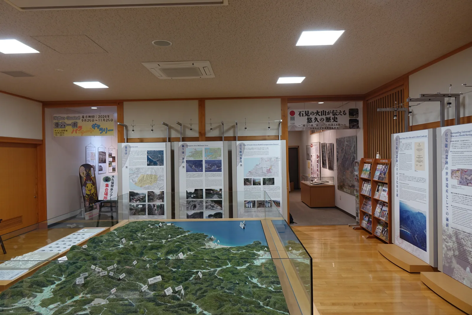 Guidance corner，前景是玻璃圍起的大型地形模型，綠色的山地延伸到藍色的海，後方立著一排解說看板與一座摺頁架，天花板是較低的白色吊頂與方形燈具