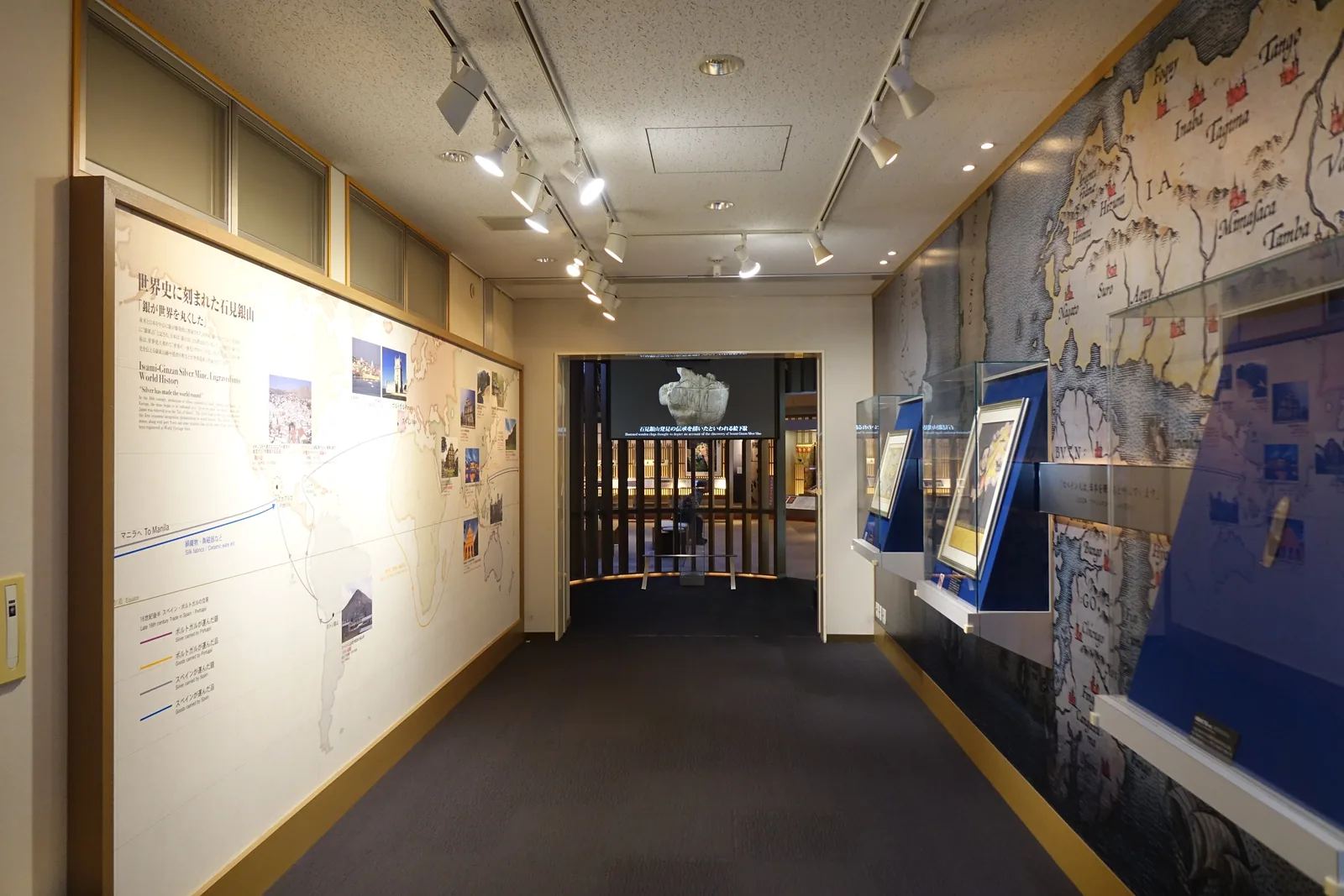 第一展示室的廊狀空間，左側牆面是世界地圖與解說，右側是古地圖與玻璃展示櫃，地面是深色地毯，天花板裝有軌道燈，盡端是一道木格柵的開口