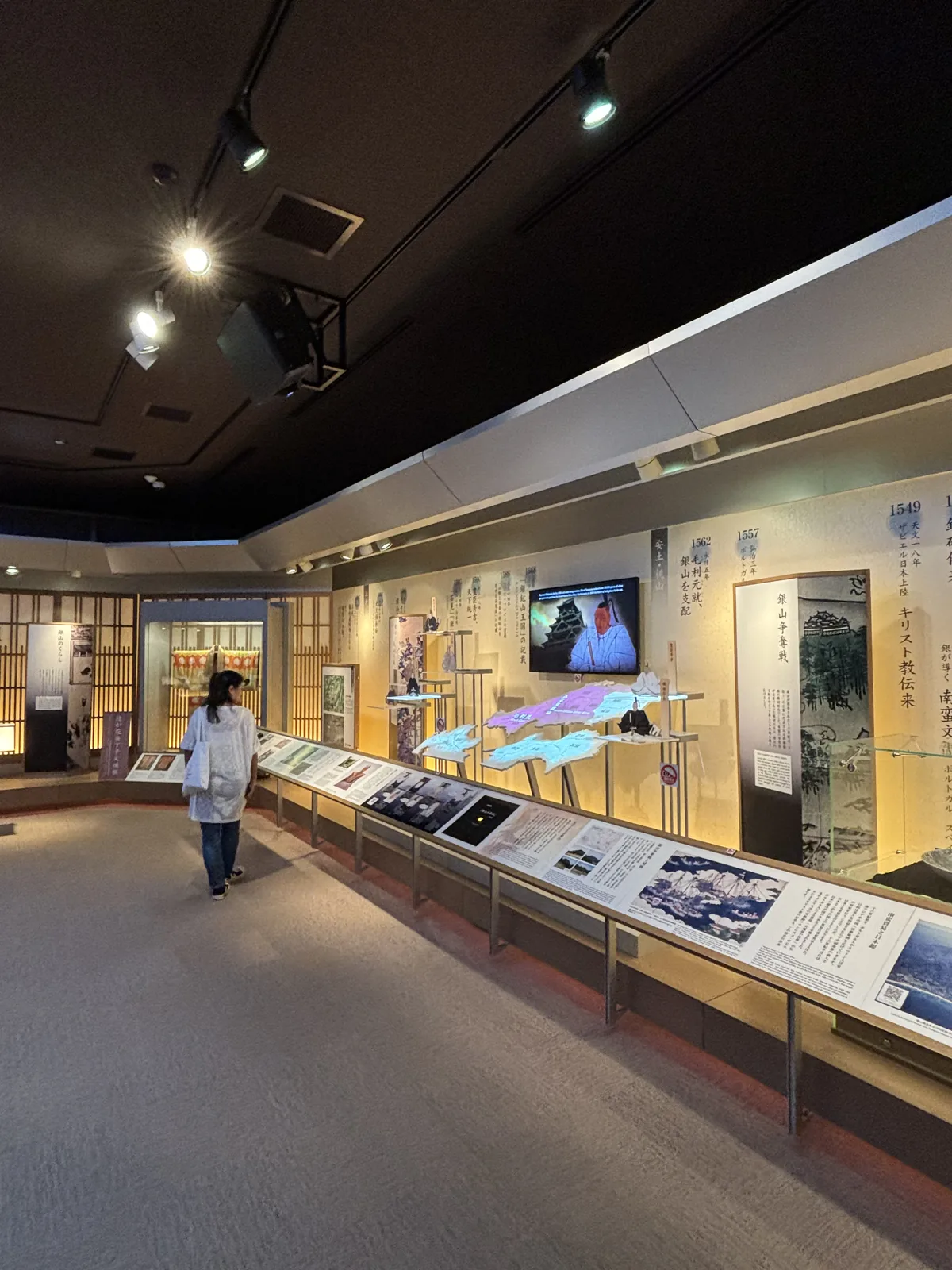 第二展示室，左側是一條連續的長形展示台與木格子分段的牆面，展示台上方掛著螢幕，天花板是深色並裝有軌道燈，一位參觀者沿展示台行走
