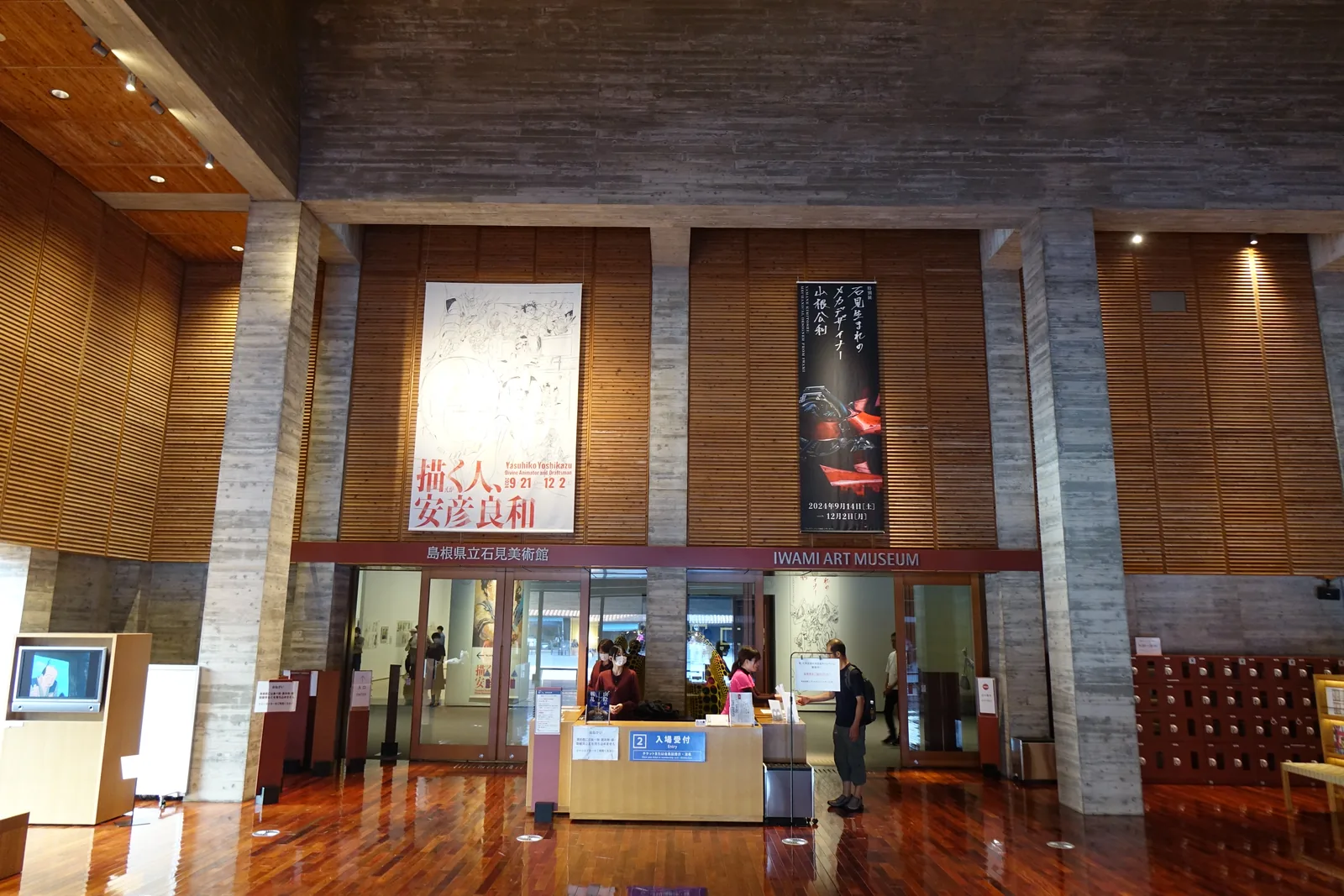 美術館展示室的入口，兩根混凝土柱之間的牆面以橫向木板覆蓋，掛著兩幅企畫展的布幔，下方是玻璃門與受理處，右側是置物櫃