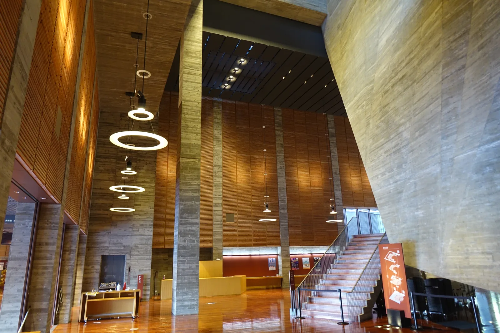 劇場大廳堂的 foyer，右側是一整面傾斜的杉板模板清水混凝土牆，左側是木板覆面的高牆與細長的混凝土柱，中央一座樓梯通往上層，天花板垂下數串圓環形吊燈